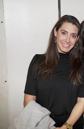 A woman with long dark hair wearing a black top and earrings, smiling at the camera, standing indoors.