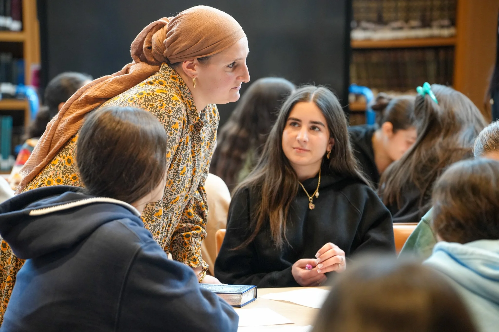 A woman wearing a beige headscarf and patterned blouse is leaning over a table, engaged in conversation with a group of students in a classroom or library setting. The students are seated around the table, and some are looking at her while others are