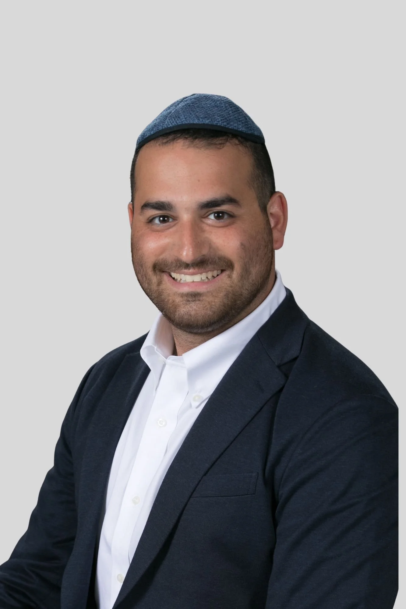 A man wearing a dark suit, white shirt, and a little blue kippah, smiling at the camera with a plain gray background.