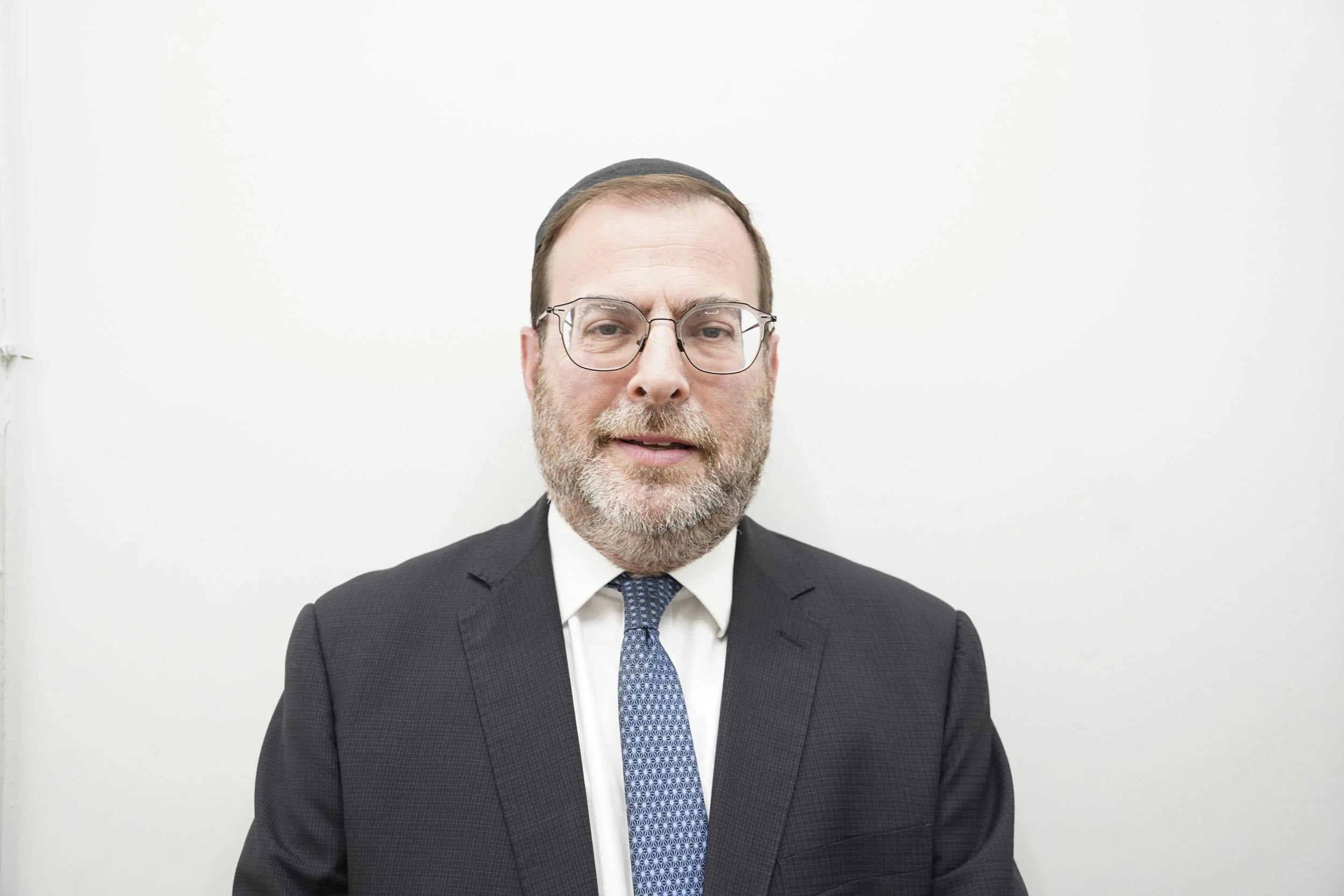 A middle-aged man with glasses, a beard, and short hair wearing a dark suit, white shirt, and a patterned blue tie, standing against a plain white wall.