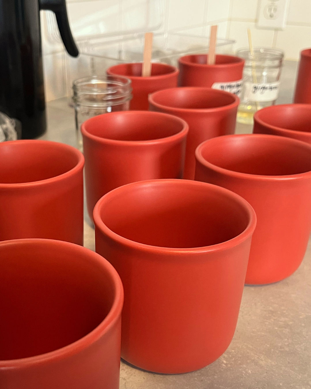 Multiple red ceramic candle jars on a kitchen counter, with some glass jars and cups in the background.