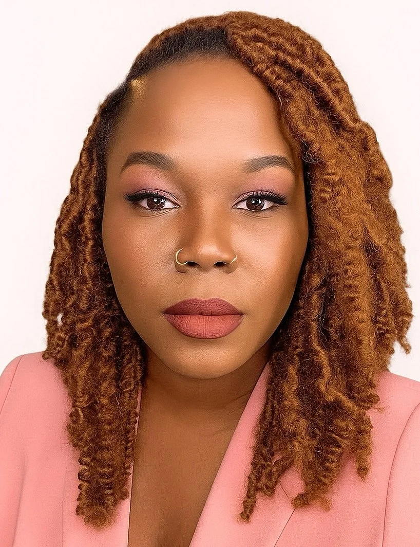 A woman with medium brown skin, reddish-brown twarled hair, and pink eyeshadow. She wears a gold nose ring and a pink blazer against a white background.