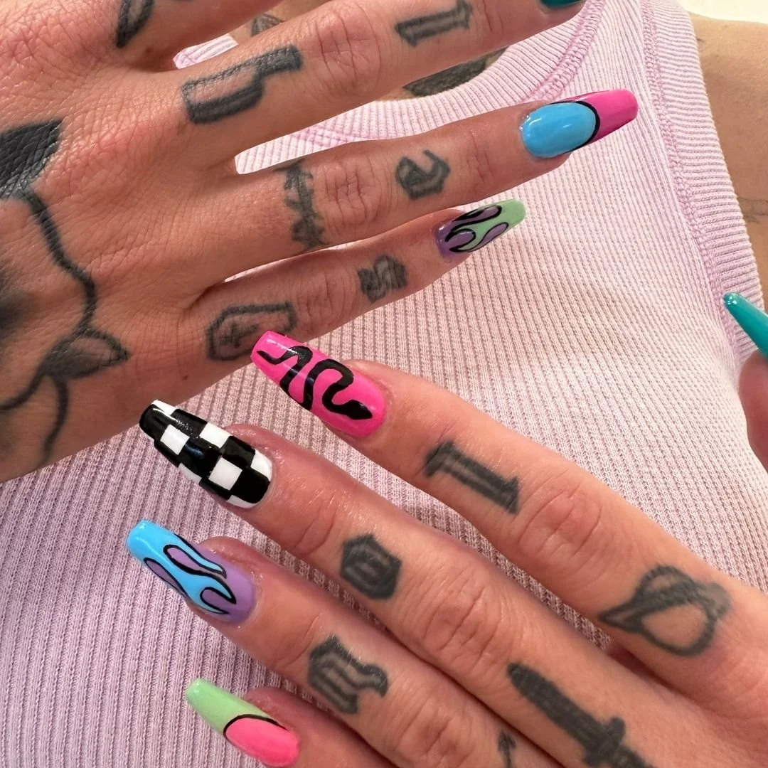 Close-up of a person's hands with colorful and patterned nail polish, tattoos, and wearing a light pink ribbed top.