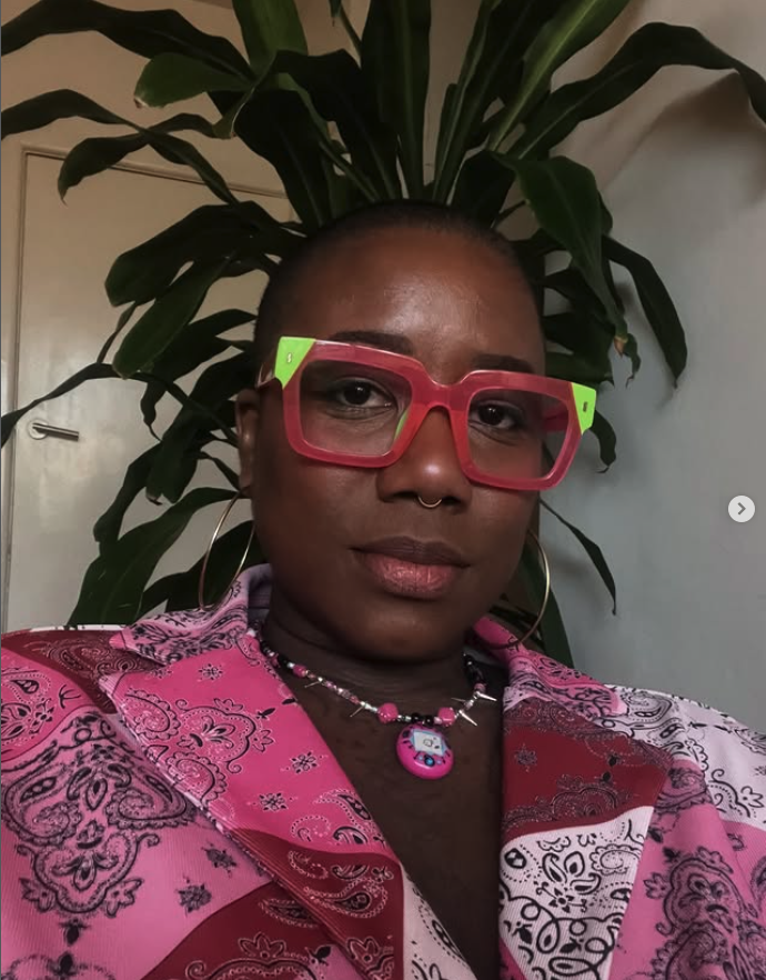 A woman with black and green hair, dark makeup, and a septum piercing, displaying tattoos of a snake and floral designs on her chest and shoulders.