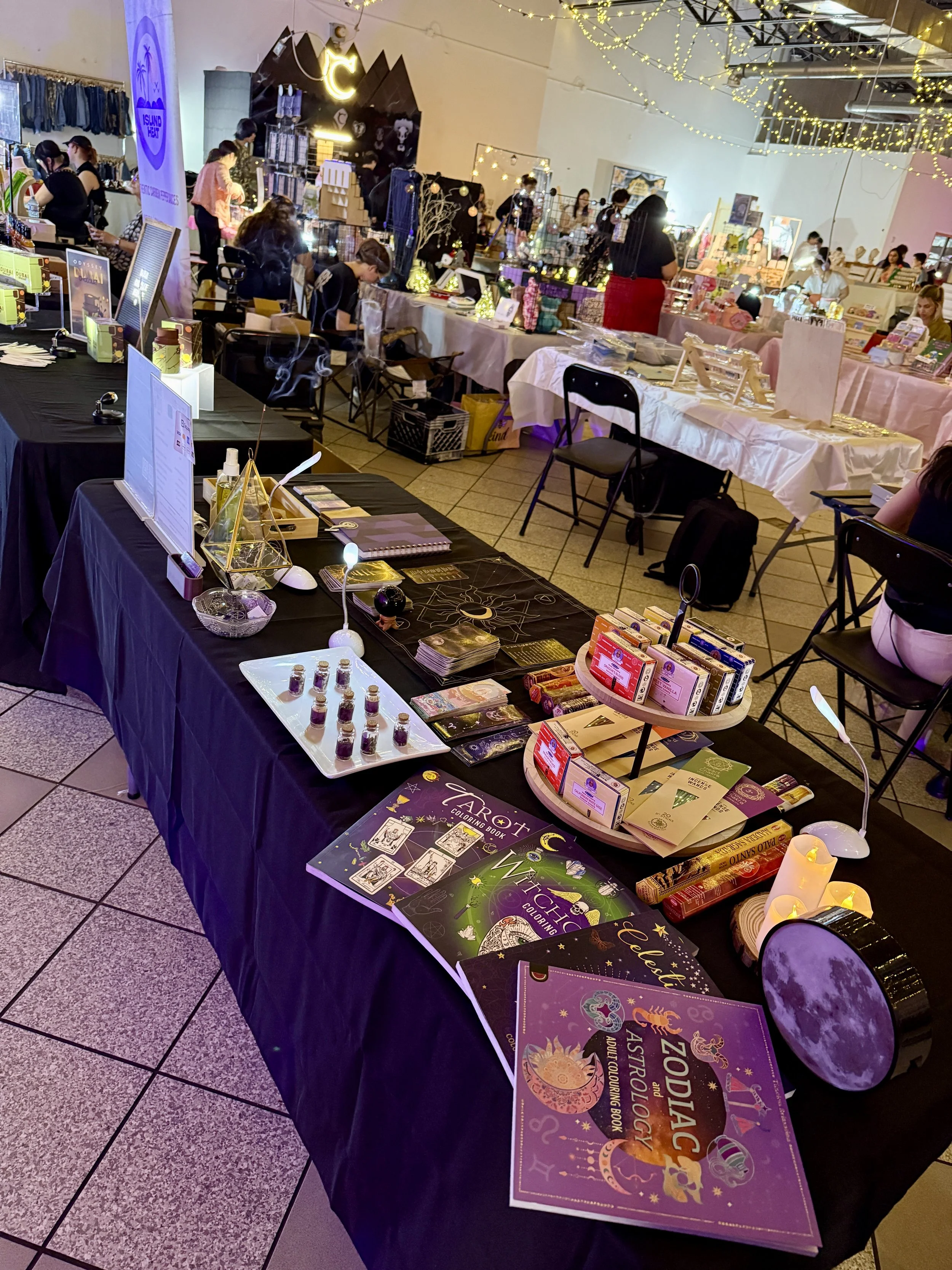Various tables set up with tarot cards, books, and crystals at a market or fair, with people browsing in the background and decorative string lights overhead.