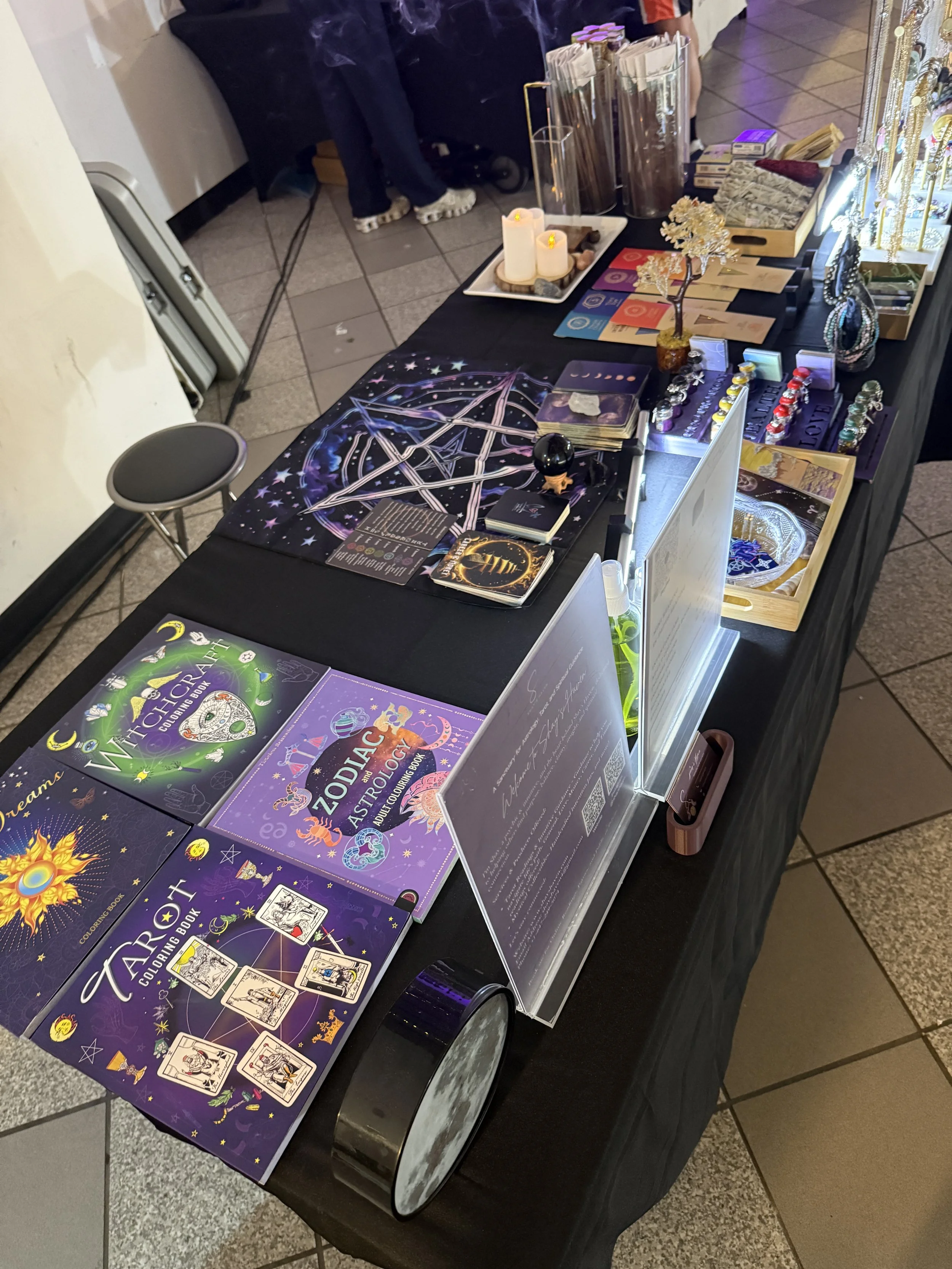 A table displaying tarot and oracle card coloring books, crystals, candles, and mystical-themed decor at a store or event.