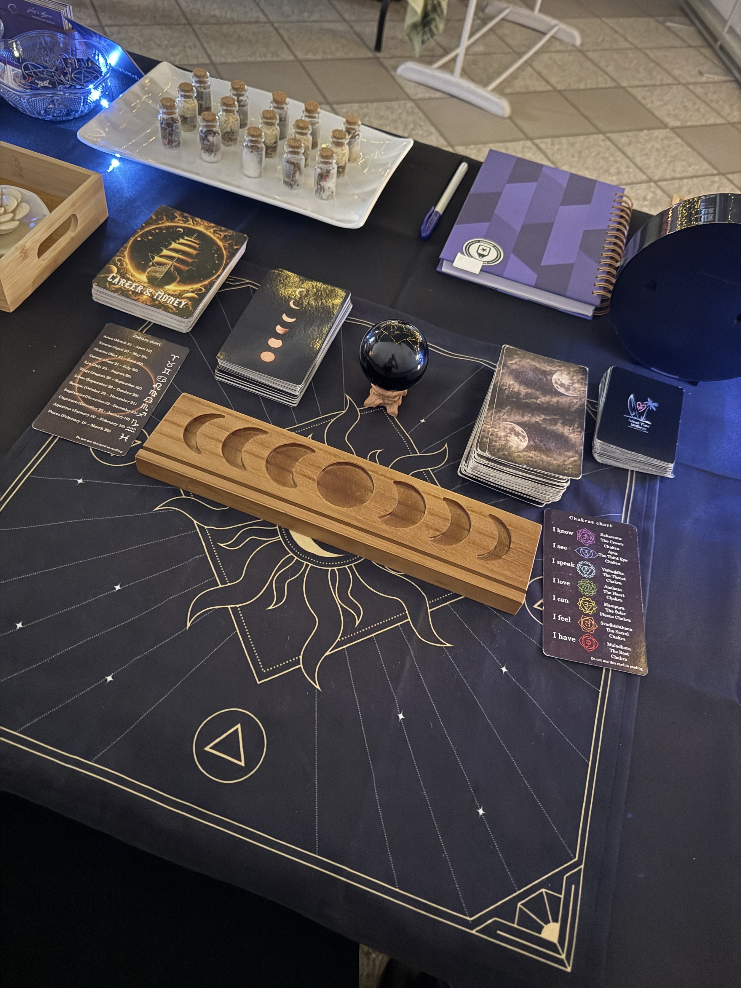 Table setup for a tarot or spiritual reading, featuring a black cloth with a gold sun design, moon phase cards, a crystal ball, chakra cards, a wooden moon phase holder, notebooks, and small bottles of herbs or crystals.