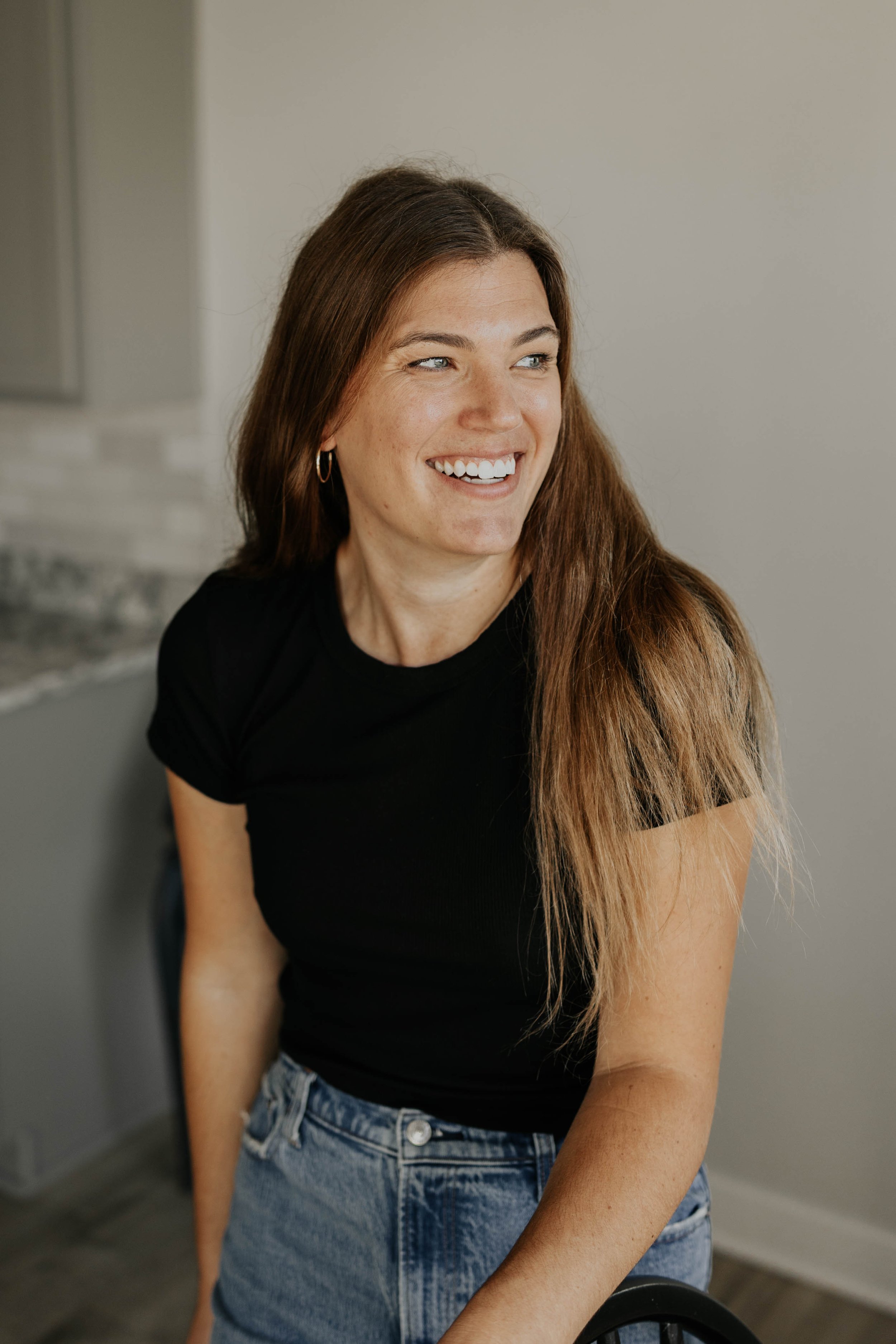 A woman with long brown hair, wearing a black t-shirt and blue jeans, smiling and looking to her right.