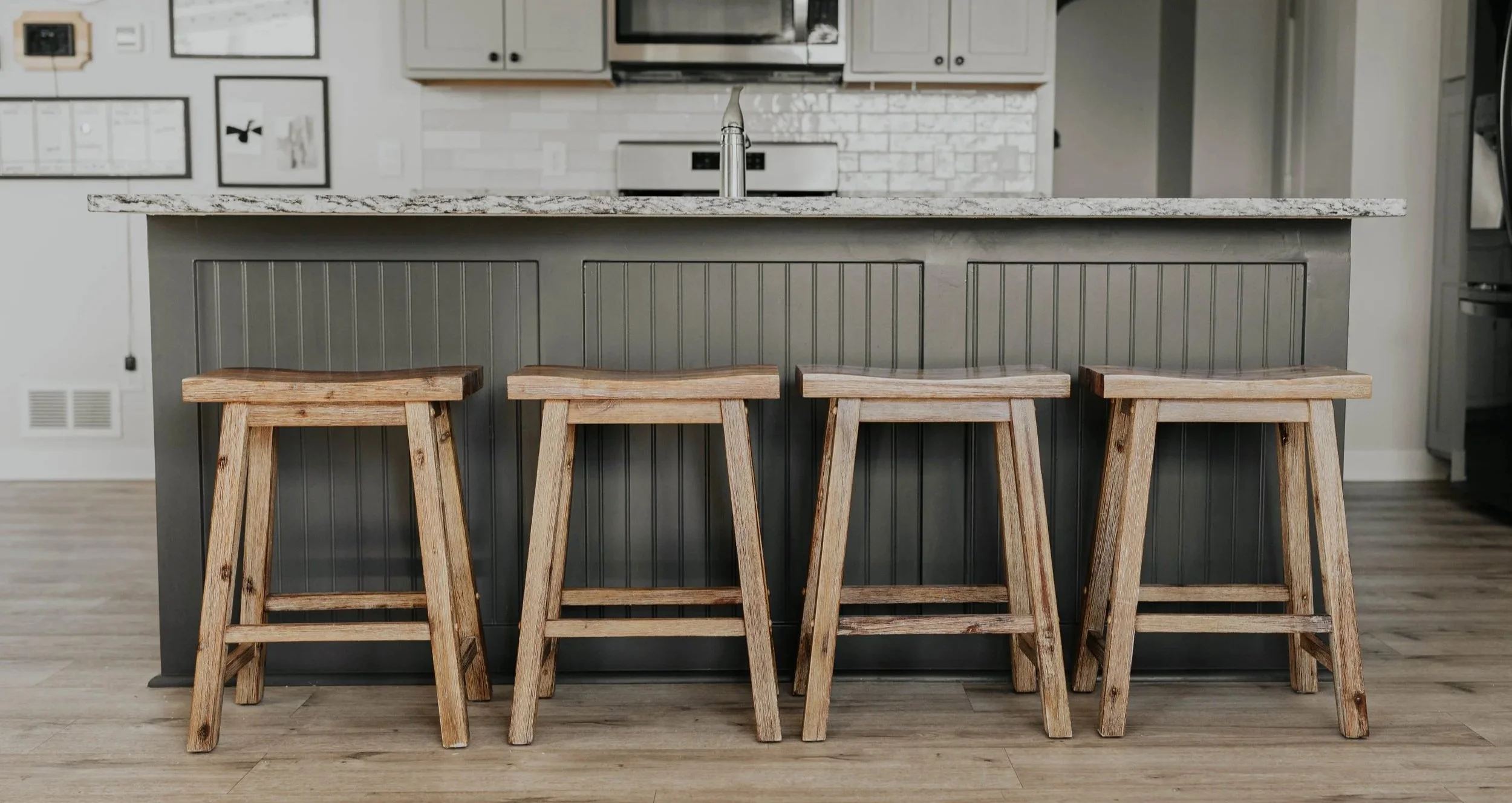 Dark gray, shiplap kitchen island with four wooden stools, gray cabinets, white zellige tile backsplash, and a granite countertop.