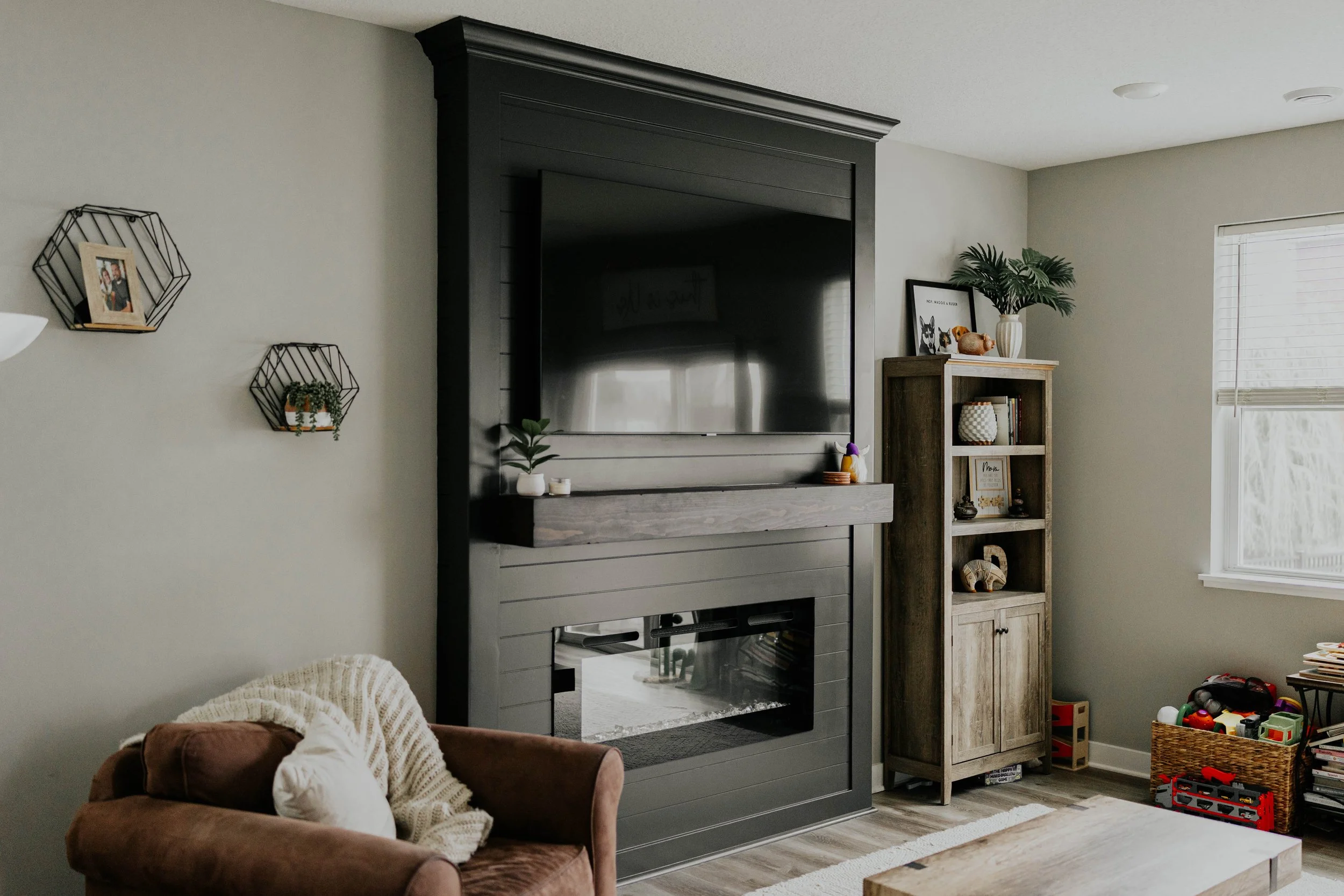 Living room with a wall-mounted flat screen TV above a modern dark gray fireplace, brown armchair with a pillow, wooden shelves with books and decorative items, and a basket of children's toys near a window.