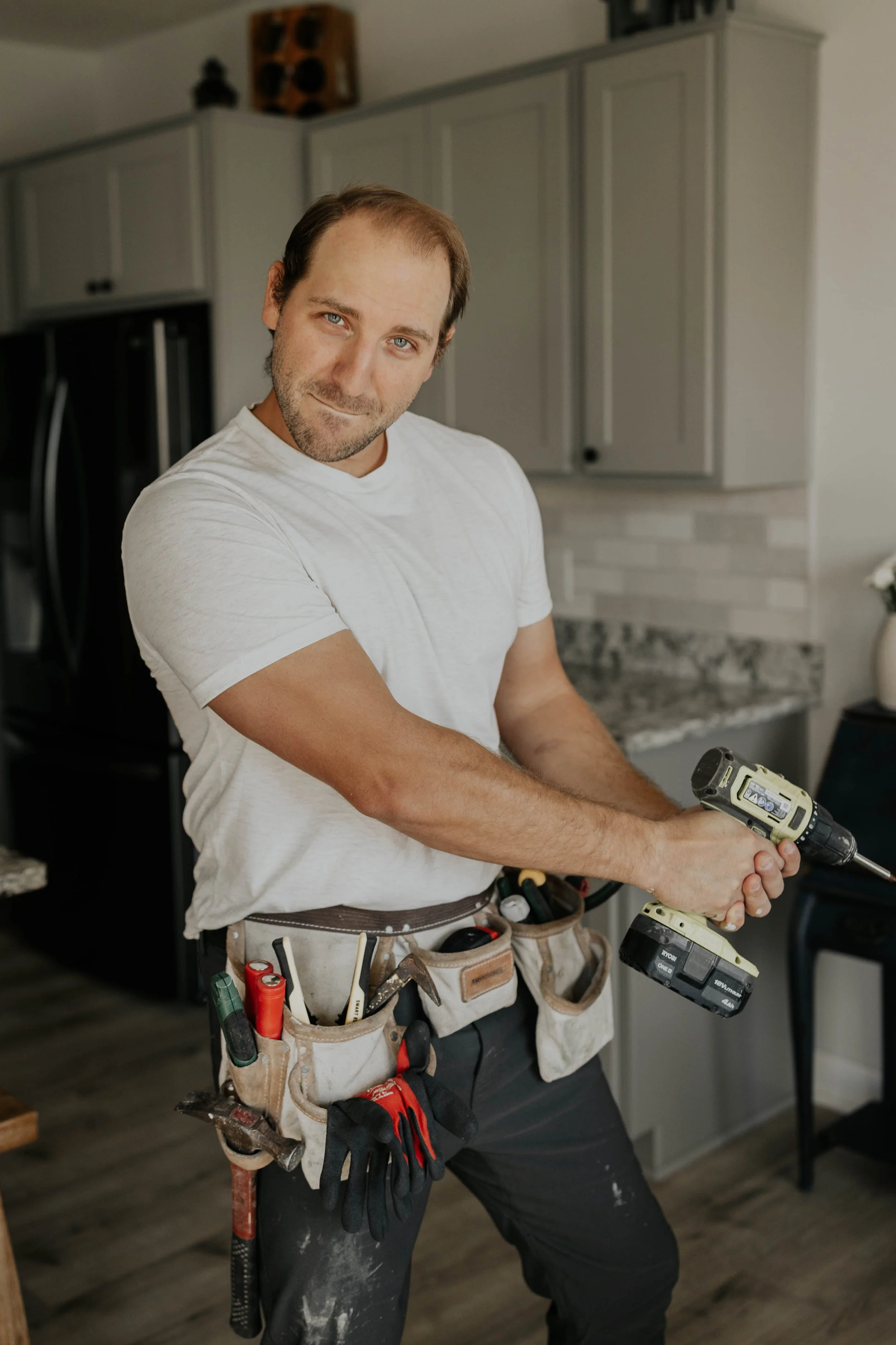 A man with a tool belt holds a cordless drill in a kitchen.