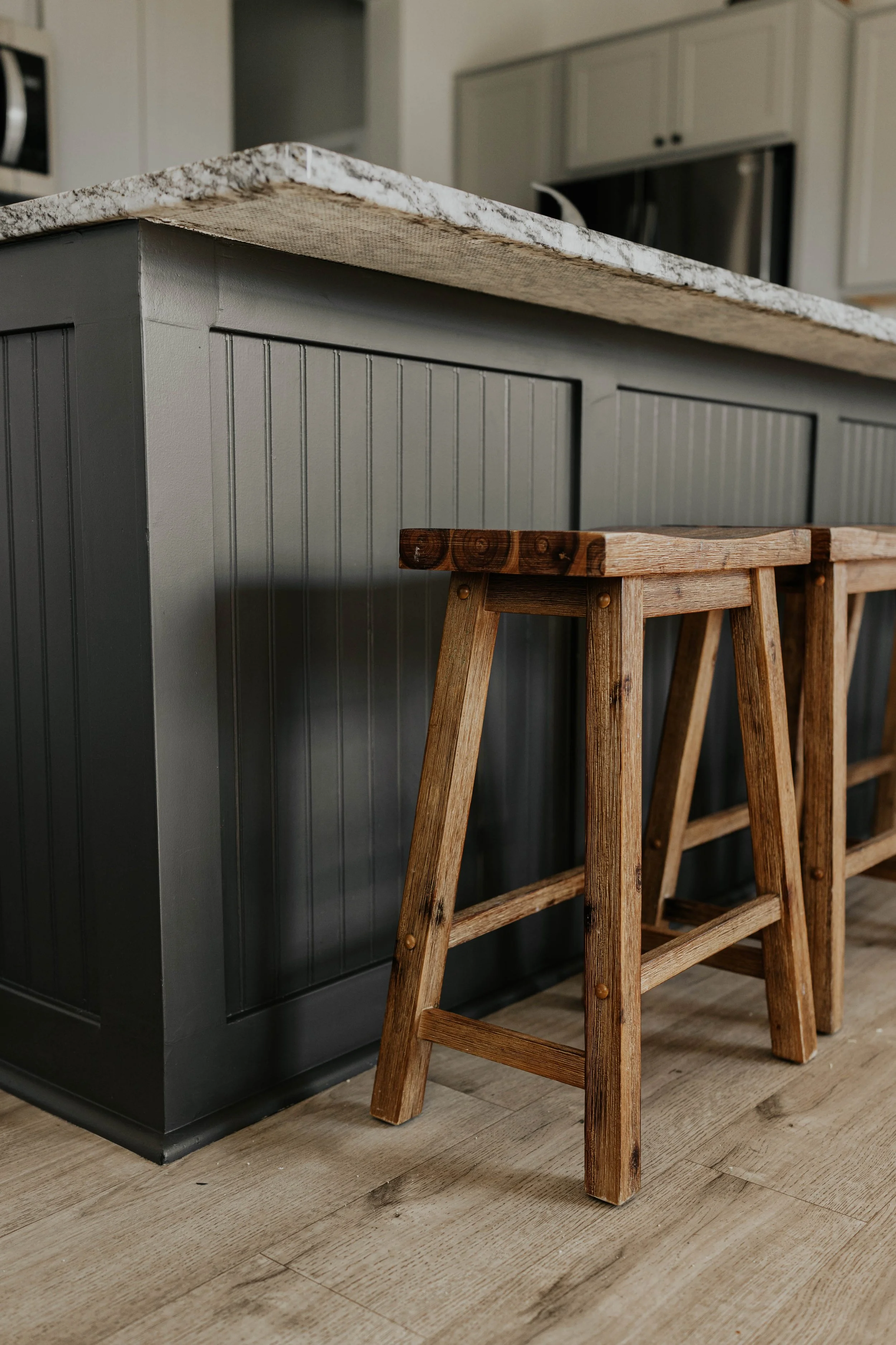 Close-up of a kitchen island with a granite countertop and gray paneled base, accompanied by wooden stools with square seats, set on a light hardwood floor.