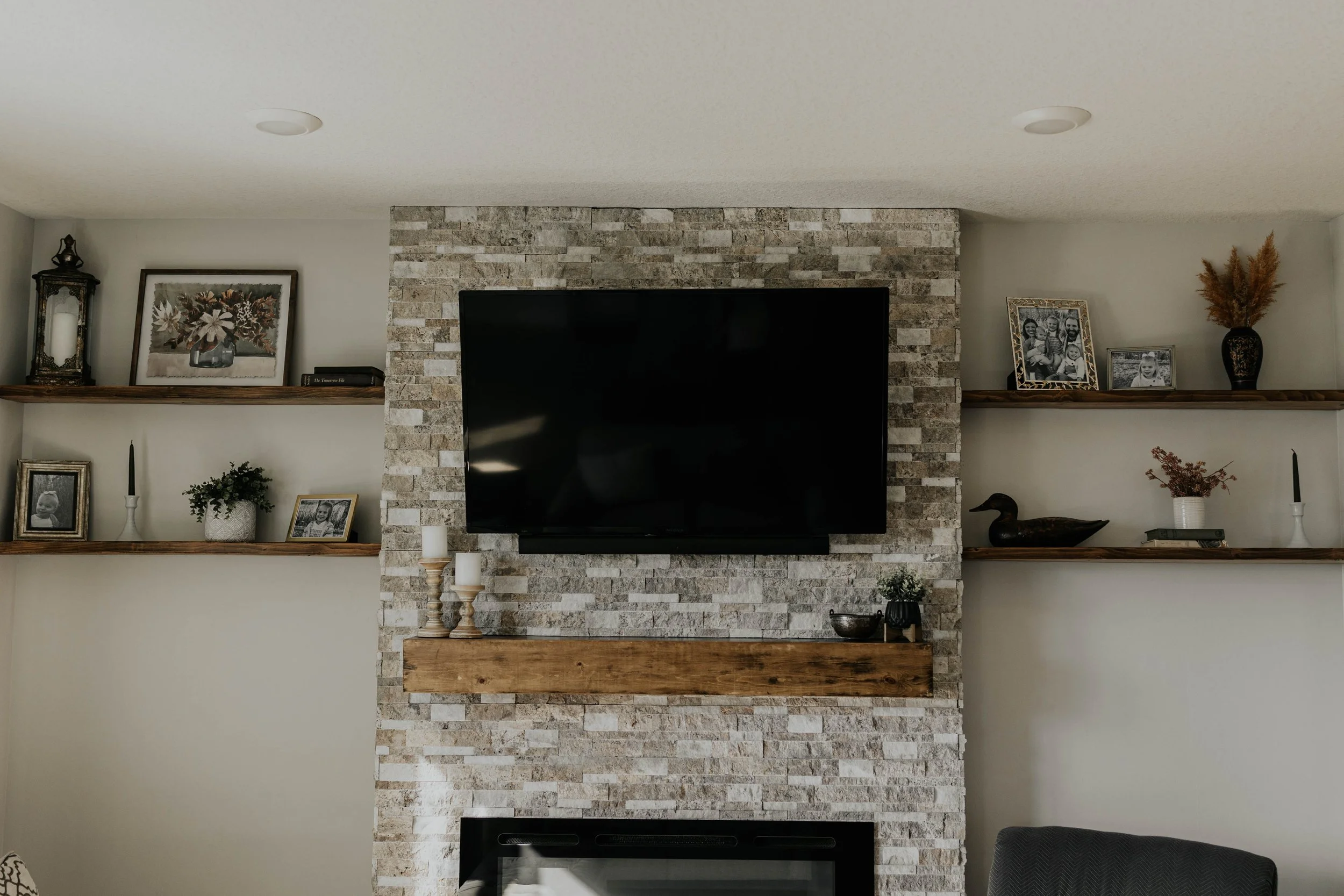Living room with a wall-mounted flat-screen TV, a stone fireplace with a wooden mantel, and floating wooden shelves holding framed photographs, potted plants, candles, a duck sculpture, and decorative items.