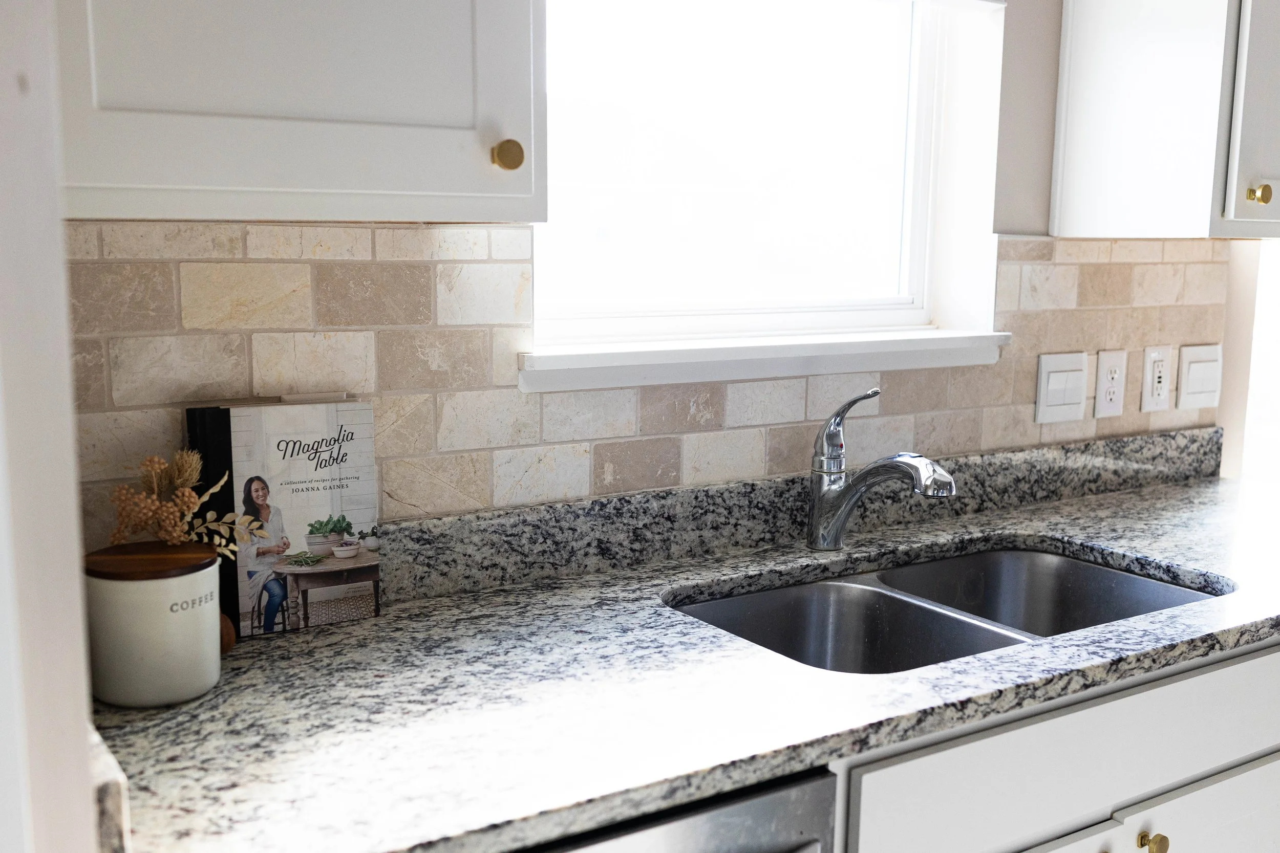Kitchen countertop with granite surface, dual stainless steel sink, chrome faucet, white cabinets with gold knobs, and a beige stone backsplash. A cookbook titled 'Magnolia Table' is on the left side of the counter, next to a coffee jar and dried dec