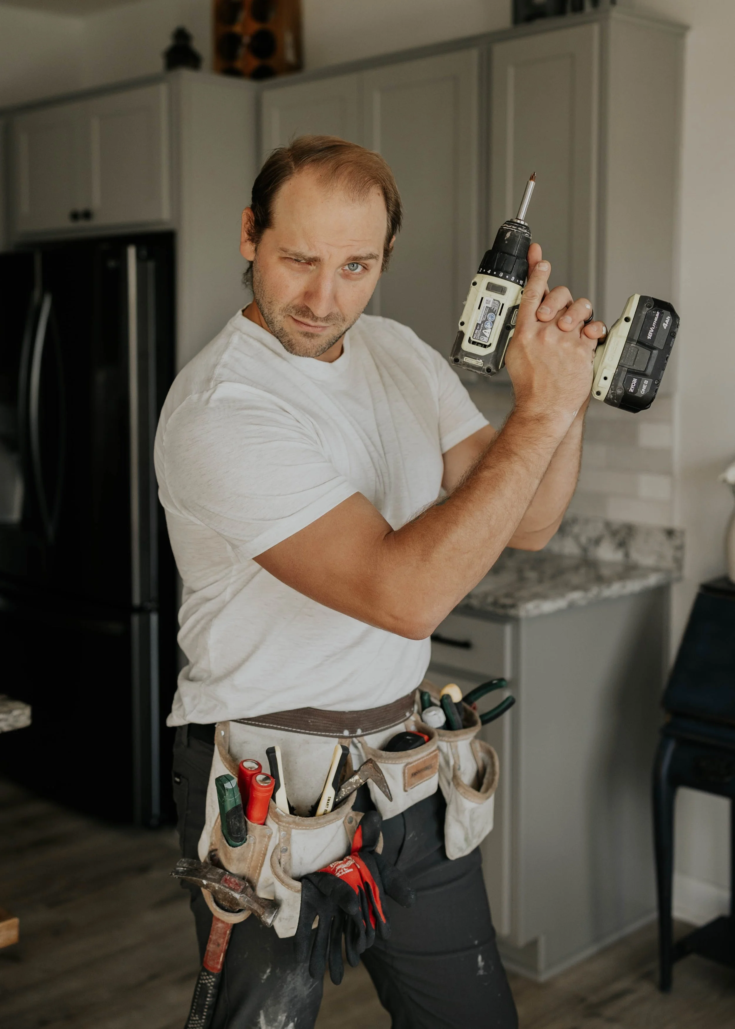 A man with a toolbox belt holding various tools, standing in a kitchen, holding a cordless drill, looking at the camera with a playful expression.