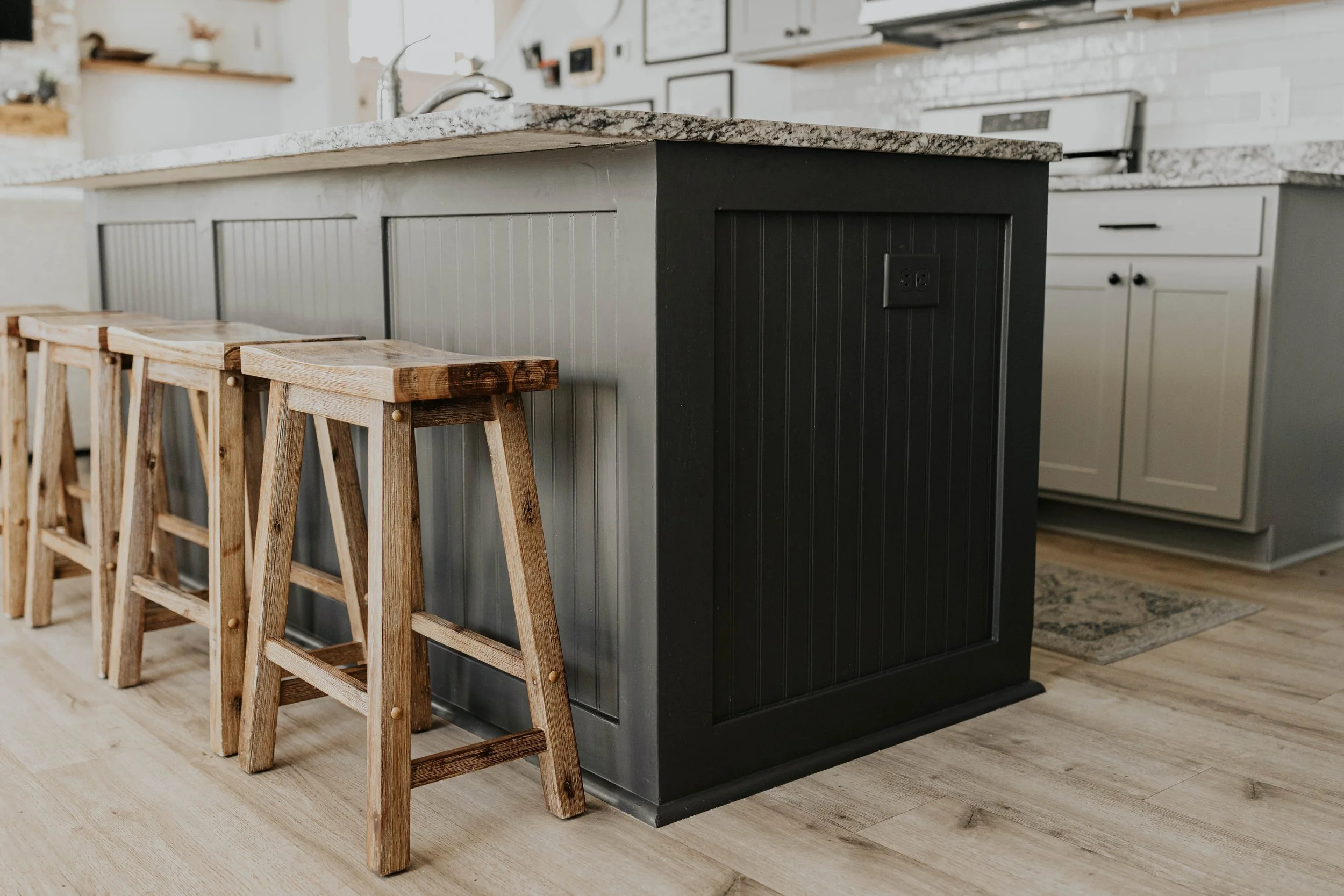 Kitchen island with a granite countertop and dark gray base, accompanied by three wooden bar stools in a modern kitchen with white cabinetry and light wood flooring.