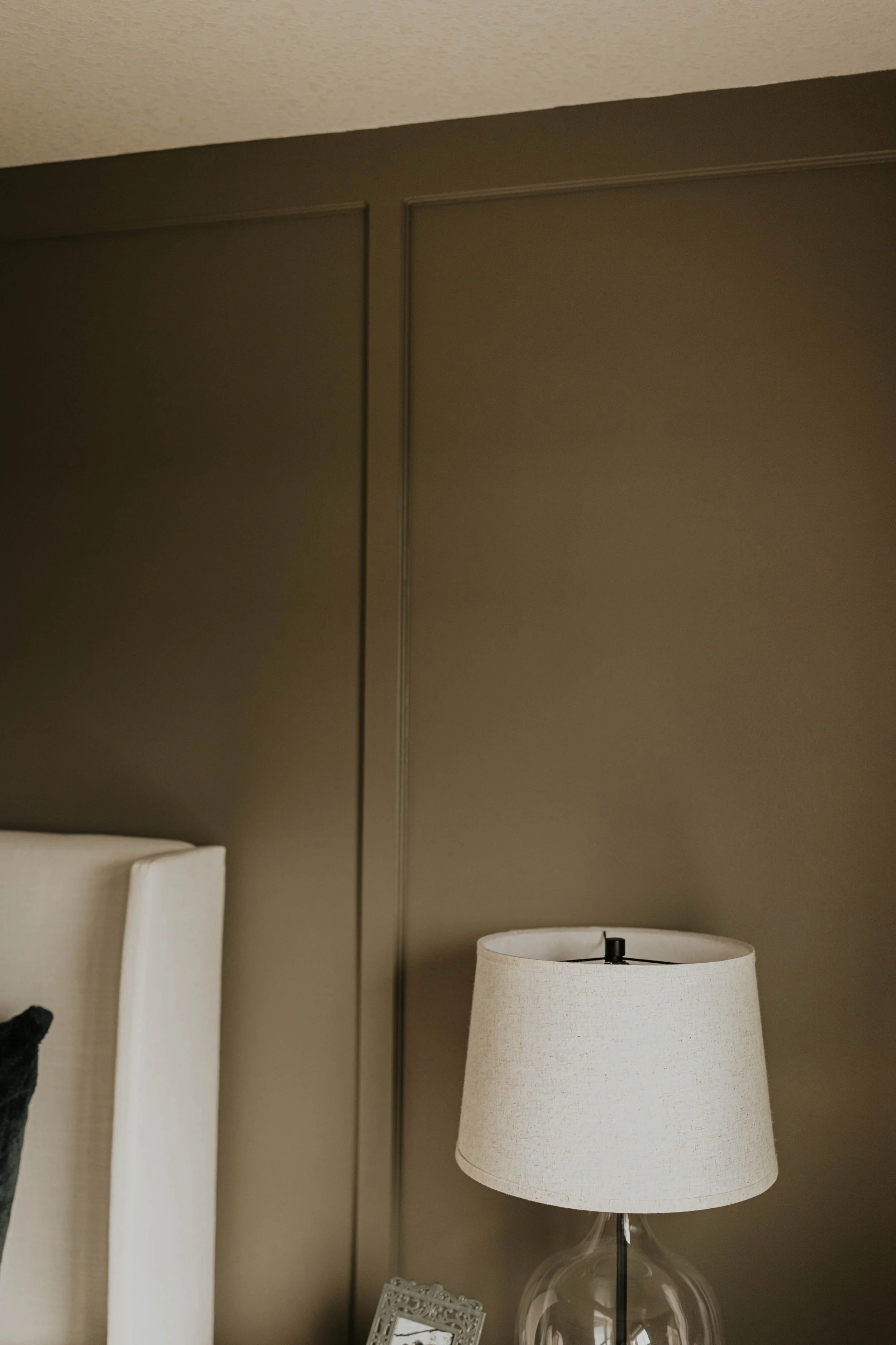 Close-up of a beige lampshade on a glass table lamp, with a dark brown board and batten wall and part of a white upholstered headboard visible in a well-lit room.