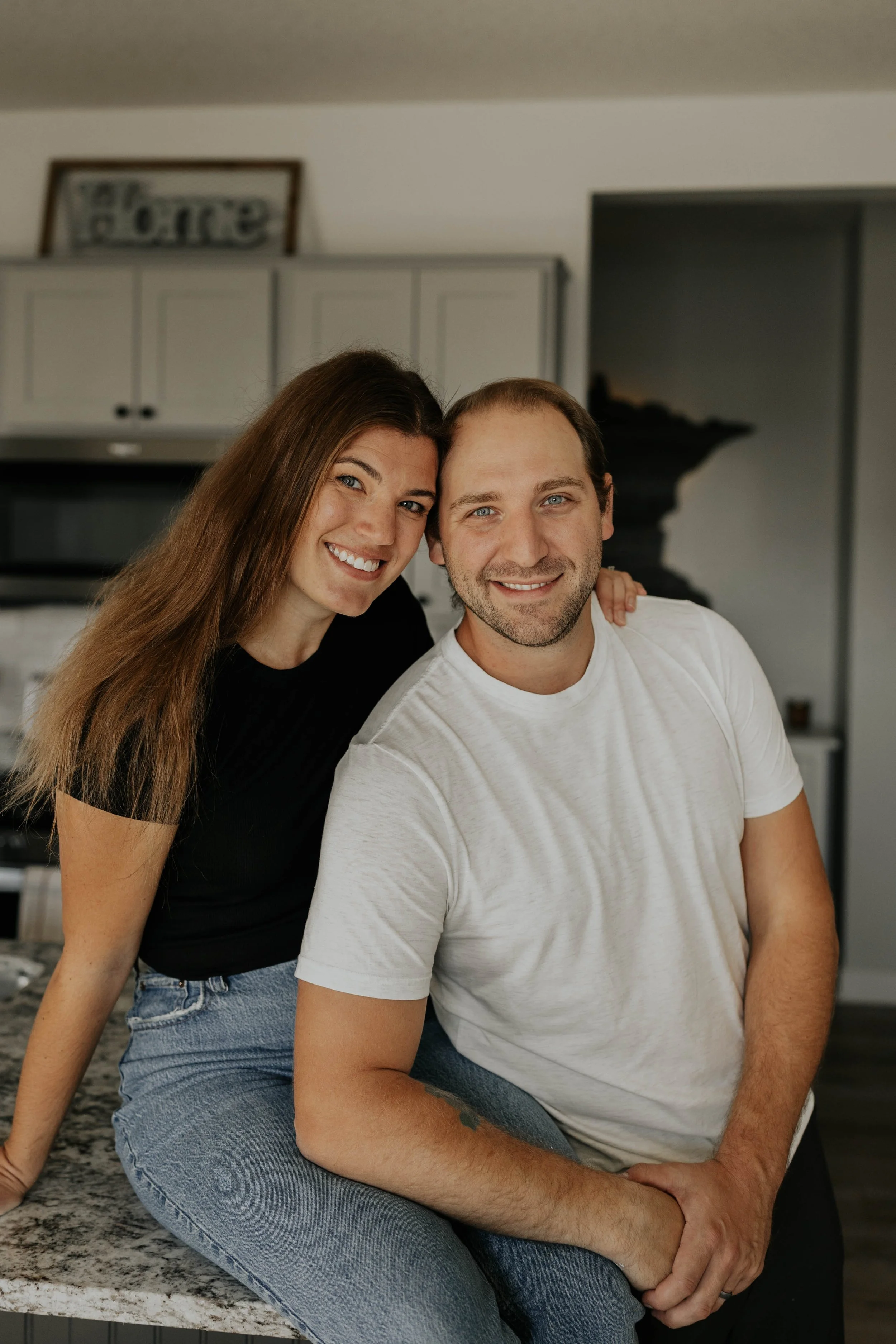 A smiling couple with the woman leaning on the man's shoulder, sitting on a kitchen countertop.