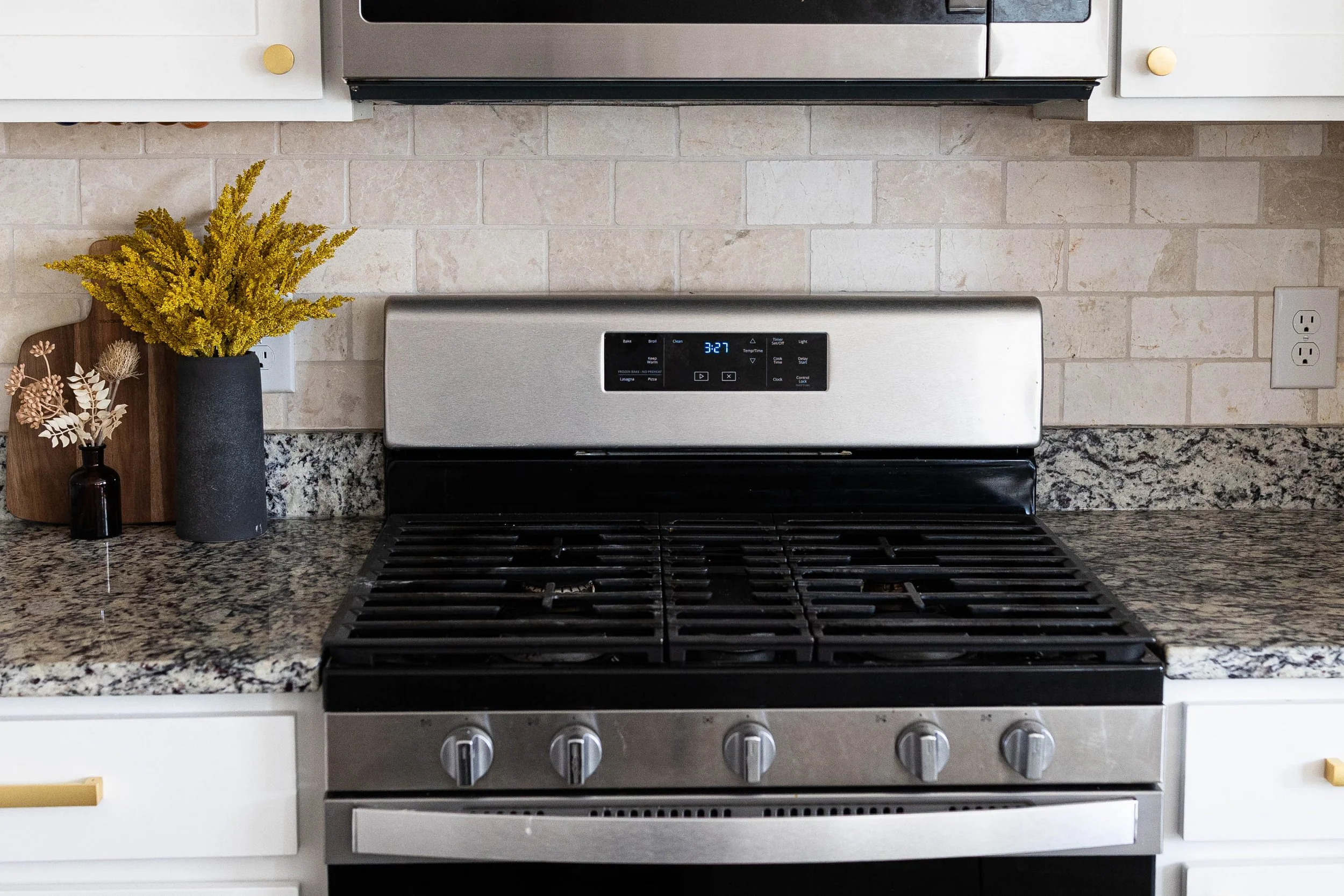 A stainless steel gas stove with five burners, black grates, and control knobs in a modern kitchen. The background features a beige brick wall, white cabinets with gold knobs, and decorative vases with plants on the granite countertop.