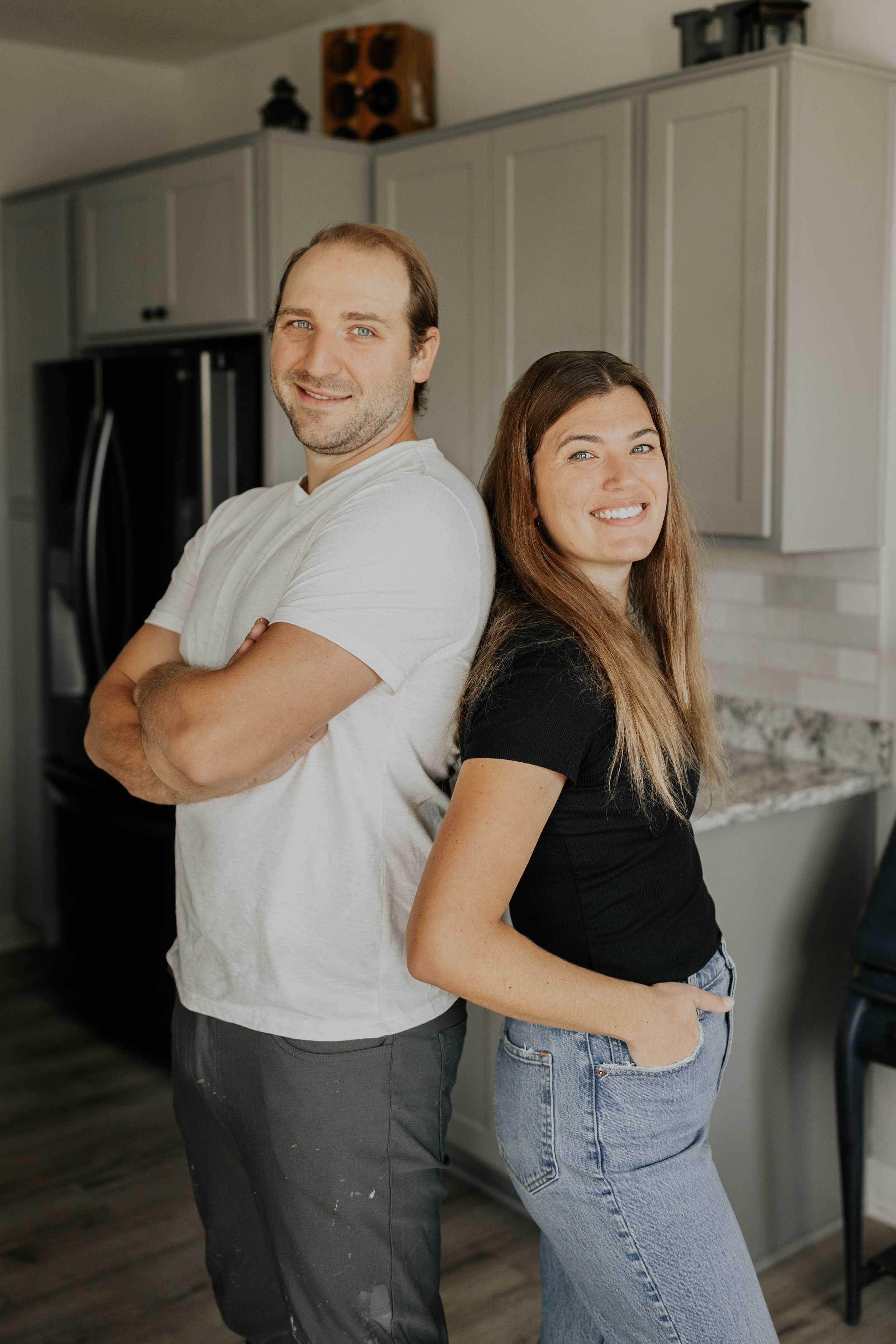 A man and a woman standing back-to-back in a kitchen, smiling at the camera.