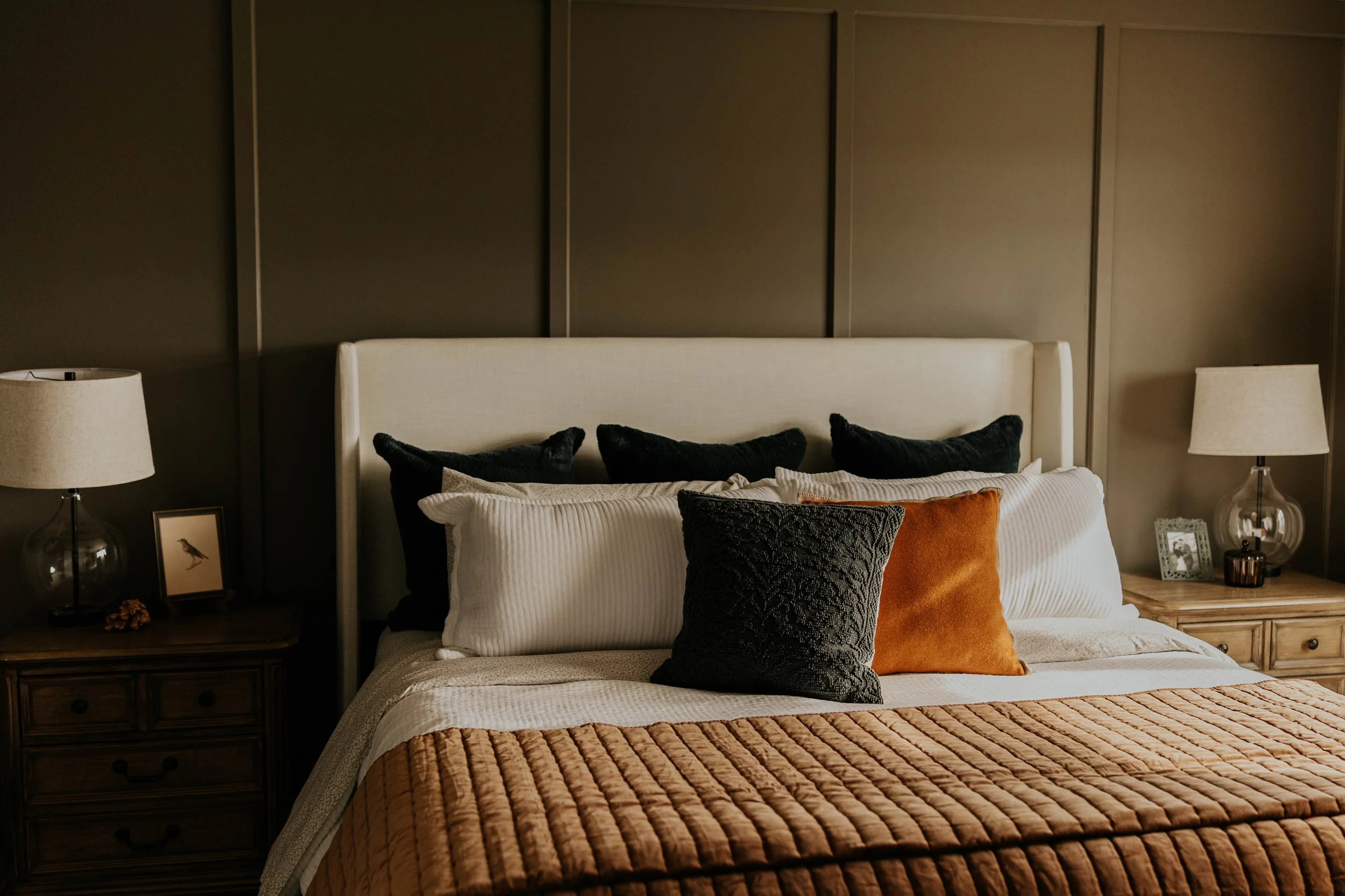 A neatly made bed with multiple pillows, including dark green, white, and camel brown colored ones, in a bedroom with dark wall panels and two nightstands with lamps and decorative items.