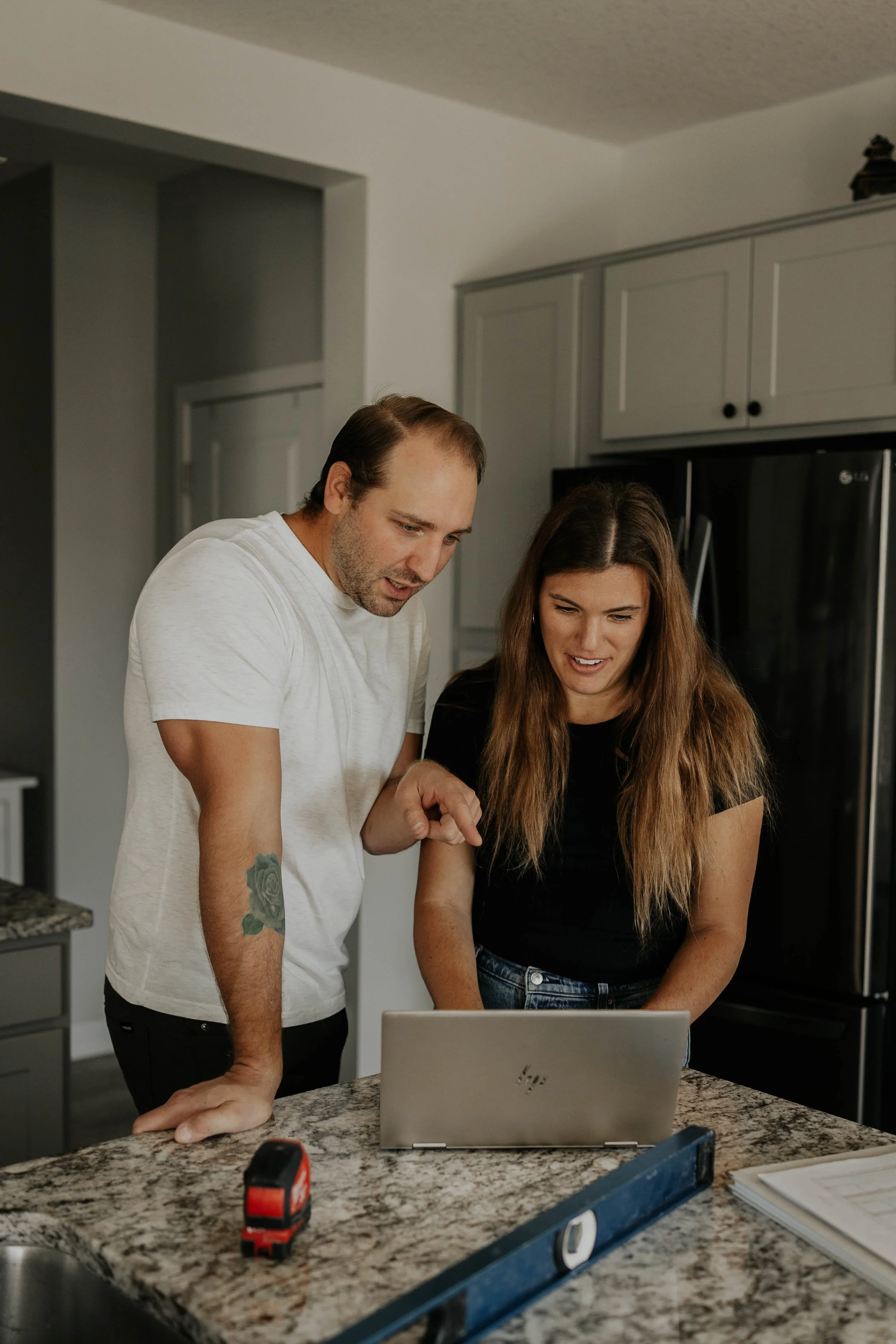 A man and woman look at a laptop on a kitchen counter, appearing to discuss something. The man has short hair and is wearing a white t-shirt, the woman has long wavy hair and is wearing a black t-shirt. There is a tape measure and a level on the counter, and the kitchen has gray cabinets and a black refrigerator.
