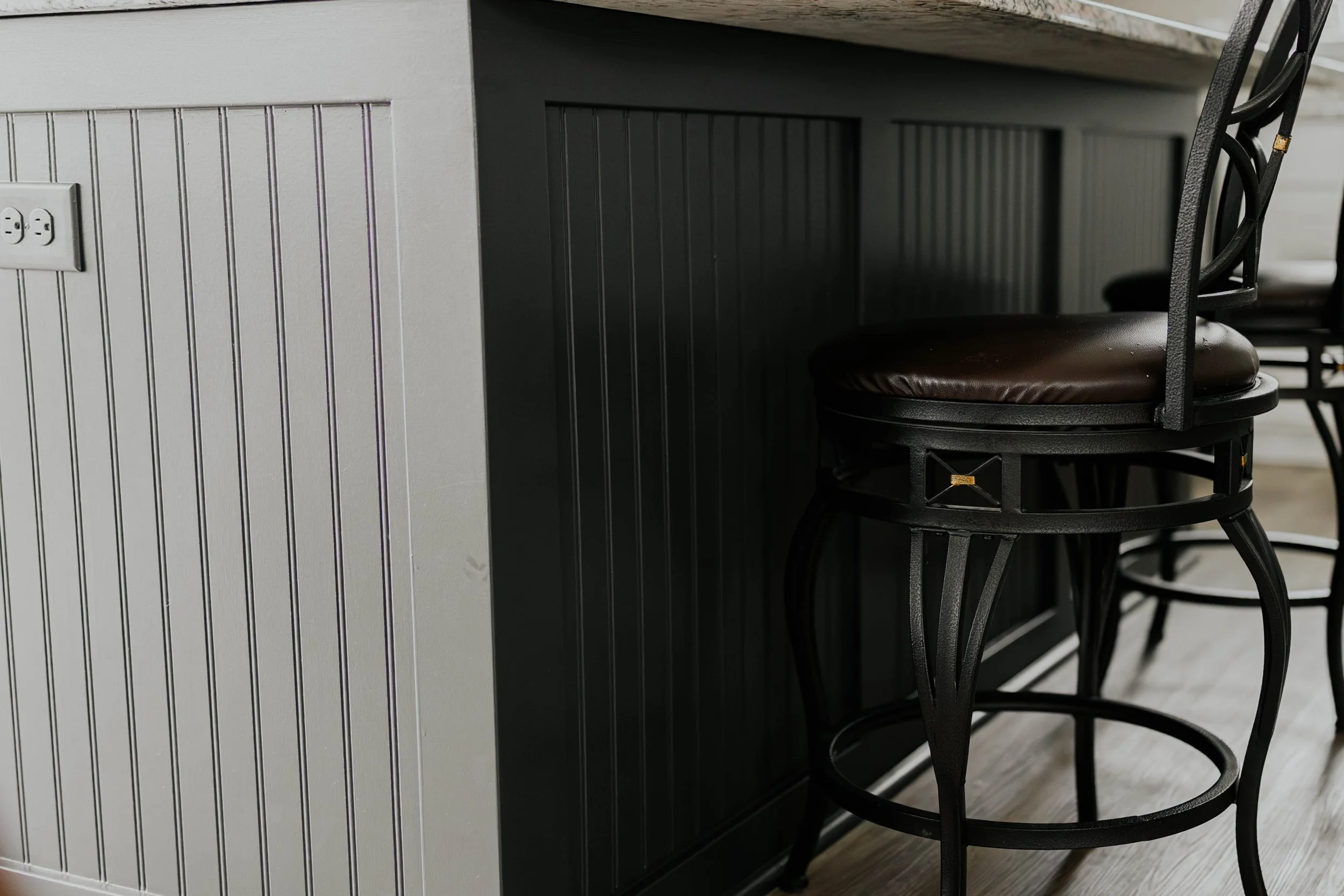 Close-up of a kitchen counter with black metal stools with brown cushioned seats, and a house electrical outlet on a white paneled wall.