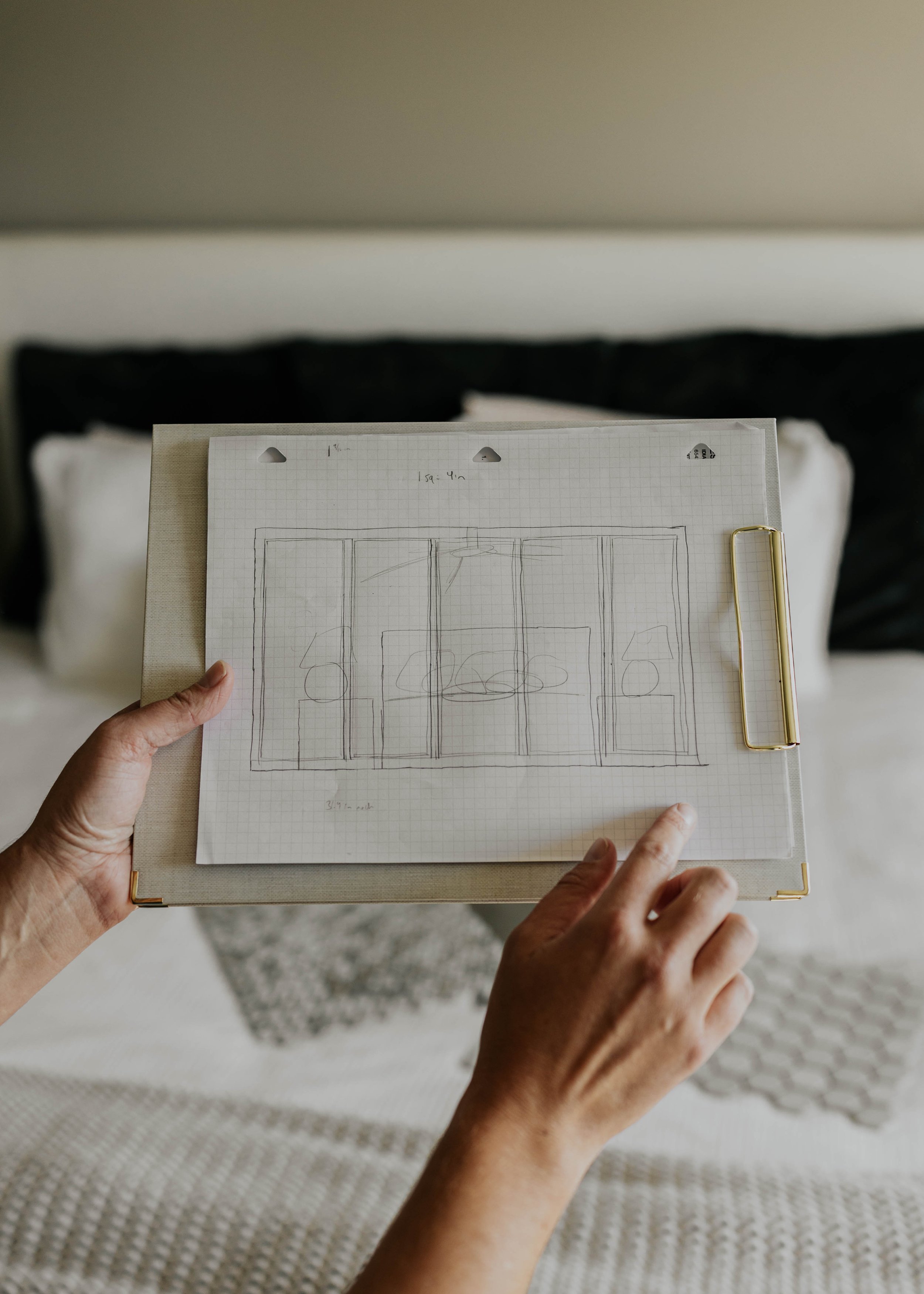 Person holding a clipboard with a hand-drawn sketch of a room layout, showing multiple sections and furniture placements, against a bedroom background.