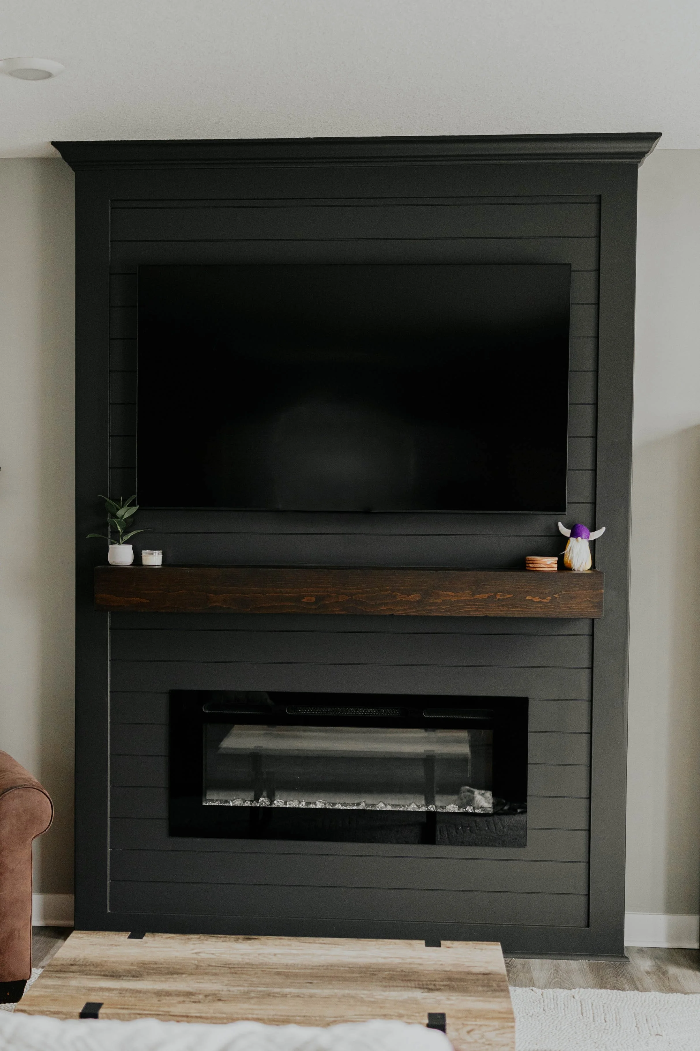 Living room fireplace with a mounted flat-screen TV above and a dark gray paneling wall, decorated with small potted plants, candles, and a novelty ornament.