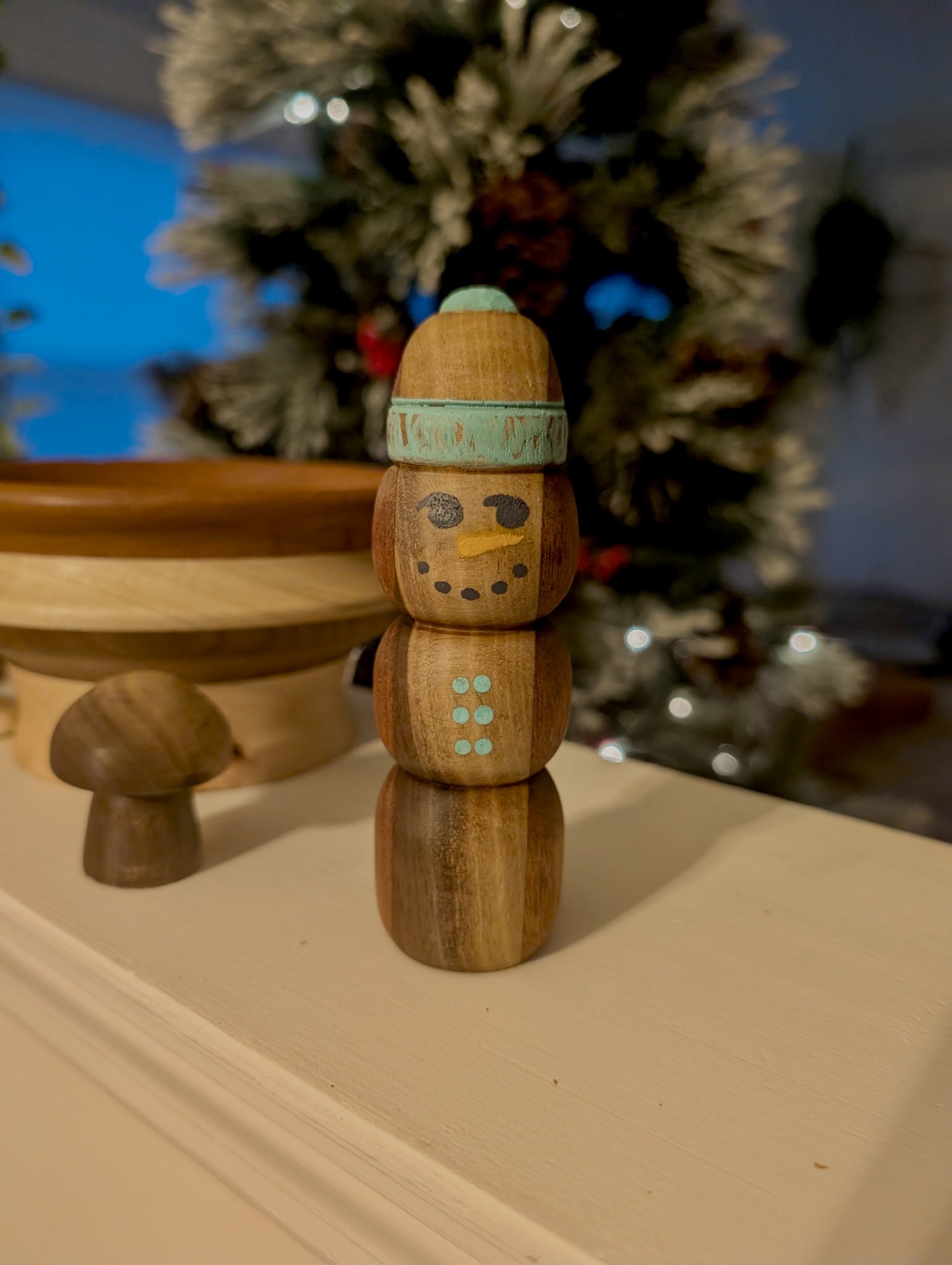 Wooden snowman figurine with painted facial features and buttons, wearing a hat, standing on a light-colored surface in front of a blurred Christmas tree with ornaments.