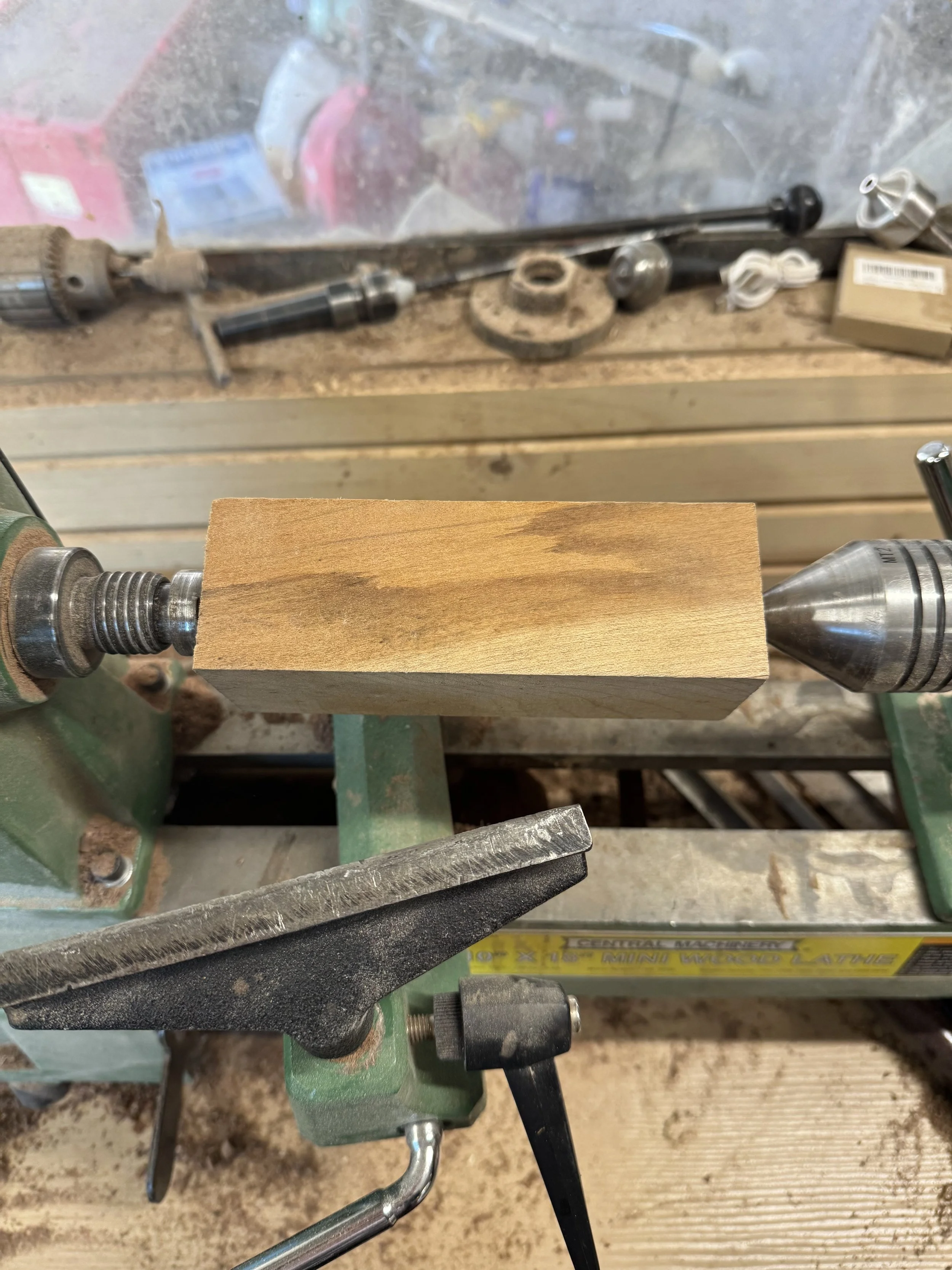 woodturning, a block of wood on a lathe in a workshop in front of a wood panel wall and a window with various lathe parts on the windowsill 