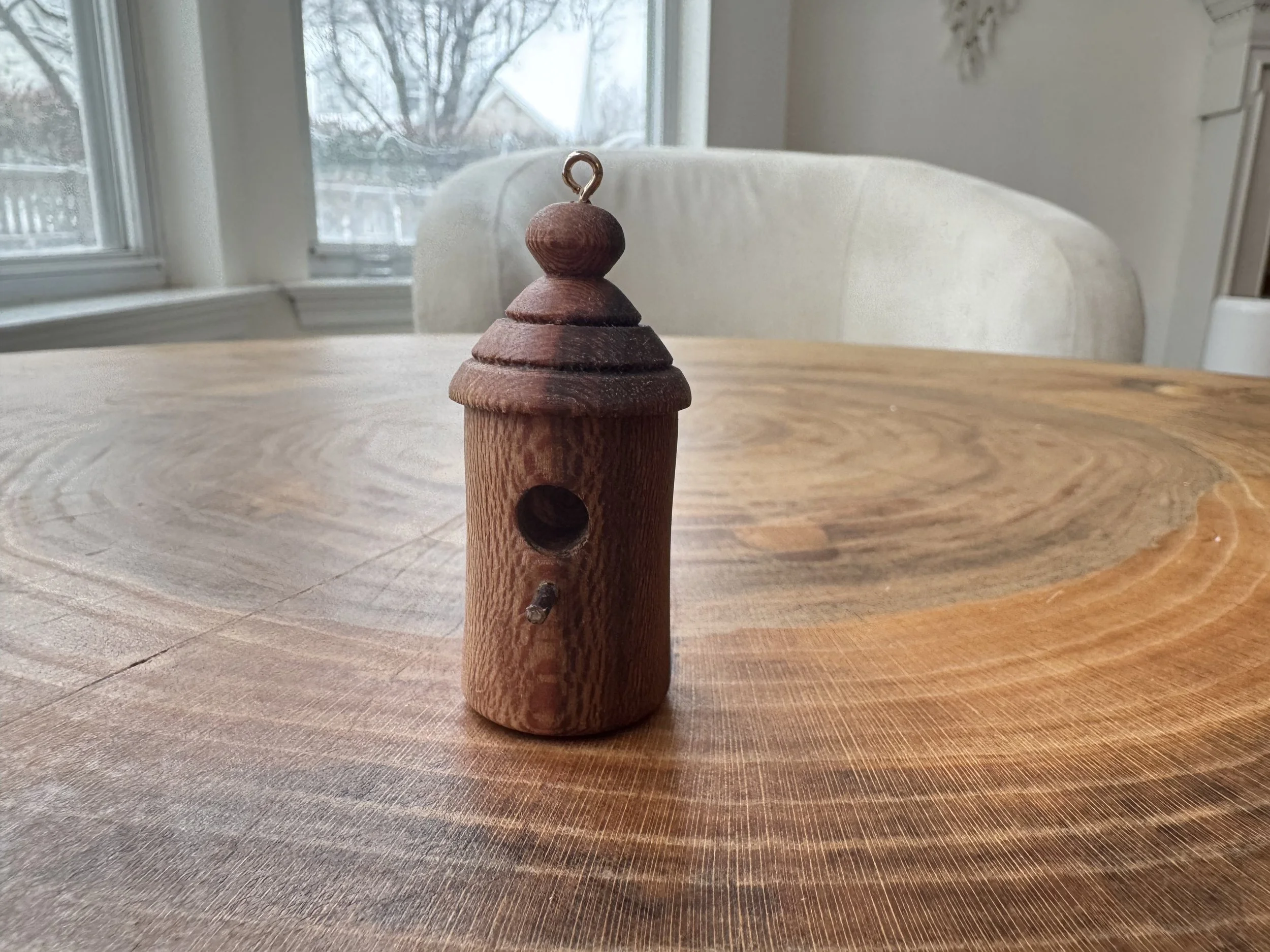 A small wooden birdhouse with a rounded, tiered roof and a circular entrance hole, placed on a wooden table inside a room with large windows and a view of trees outside.
