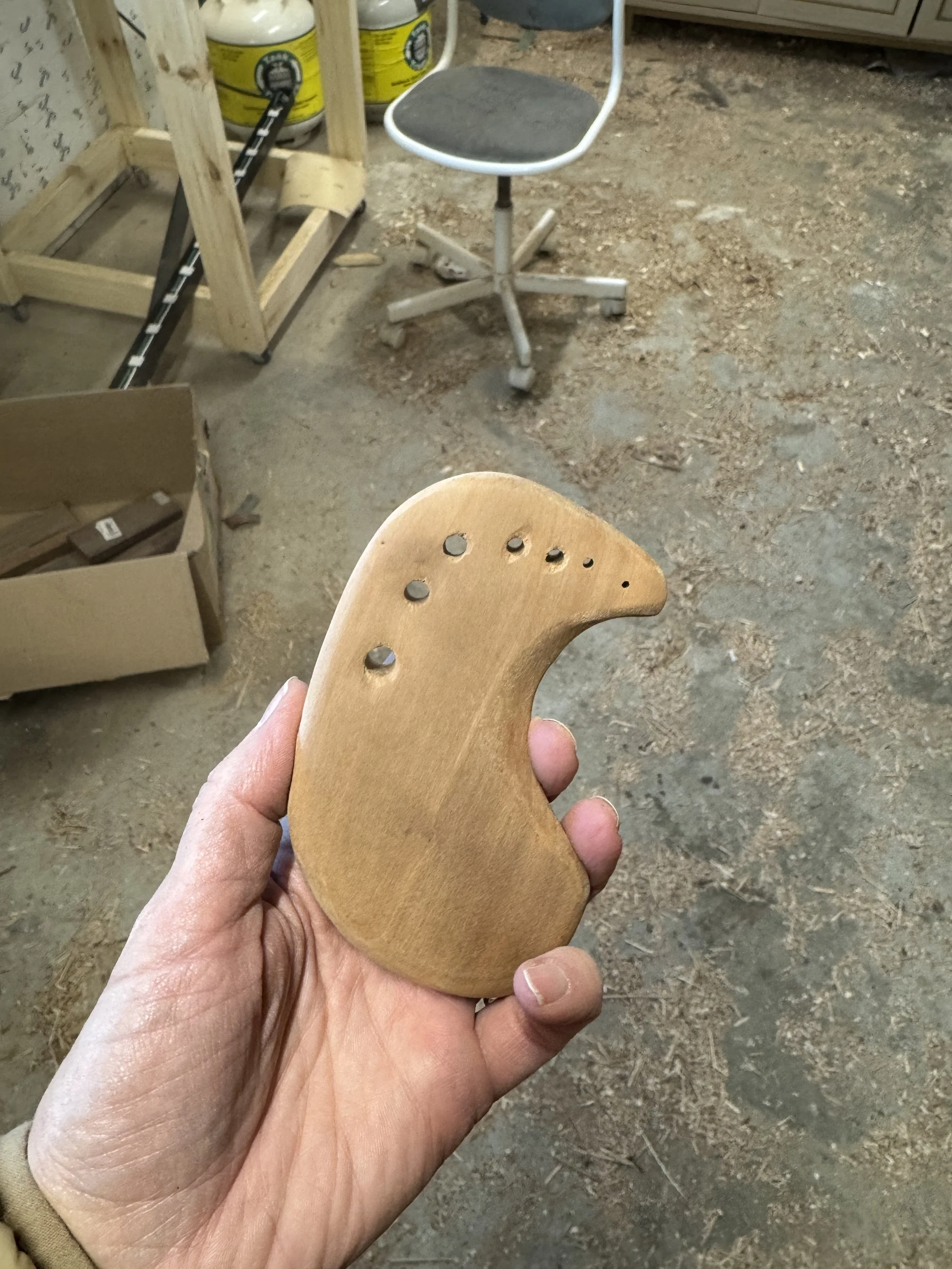 A person holding a handmade wooden herb stripper or herb puller, in what appears to be a woodworking workshop with tools, a chair, and wooden materials in the background.