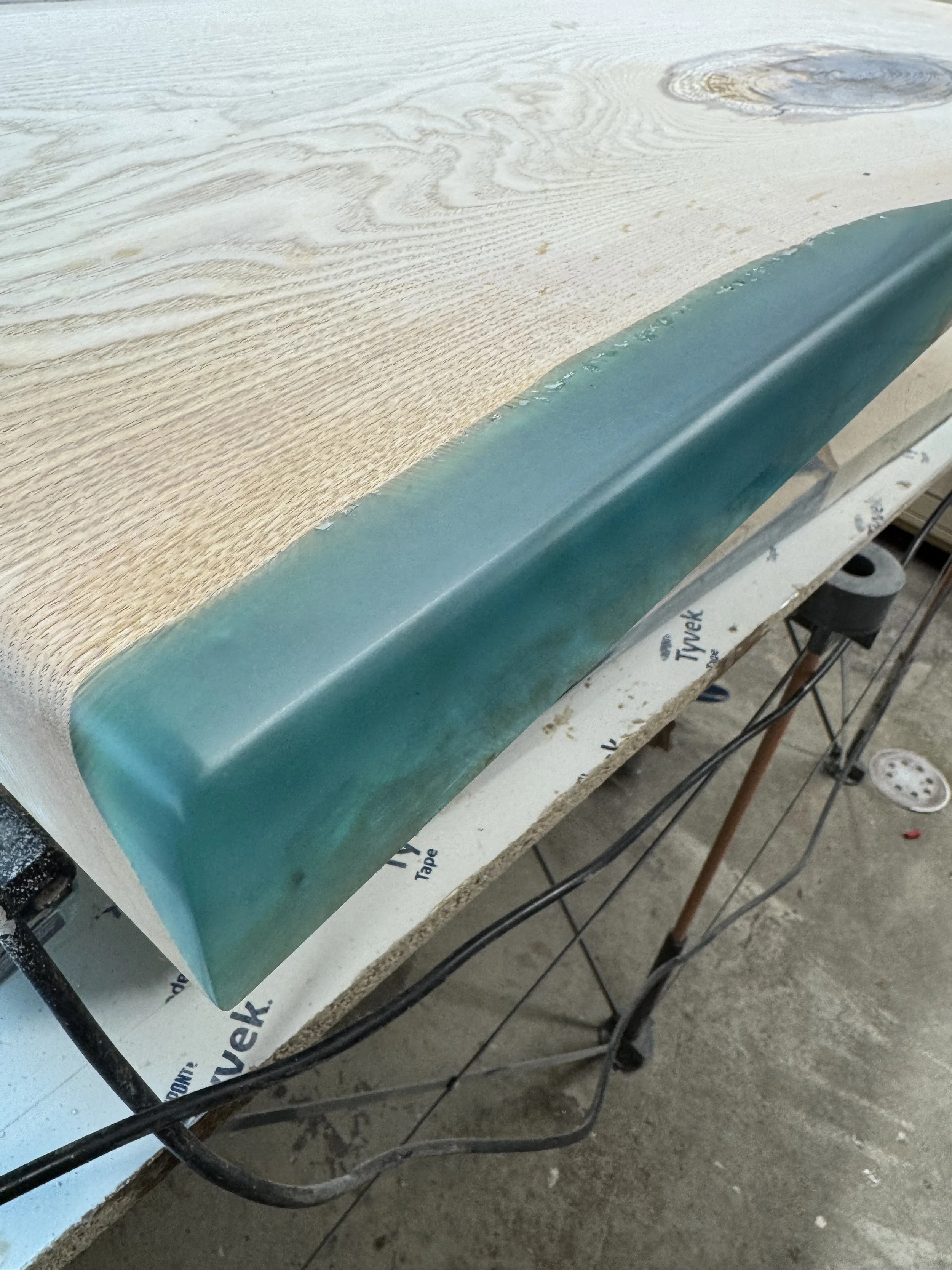 Close-up of a wood table with a teal-colored corner edge, resting on a worktable with tools and workshop items visible.