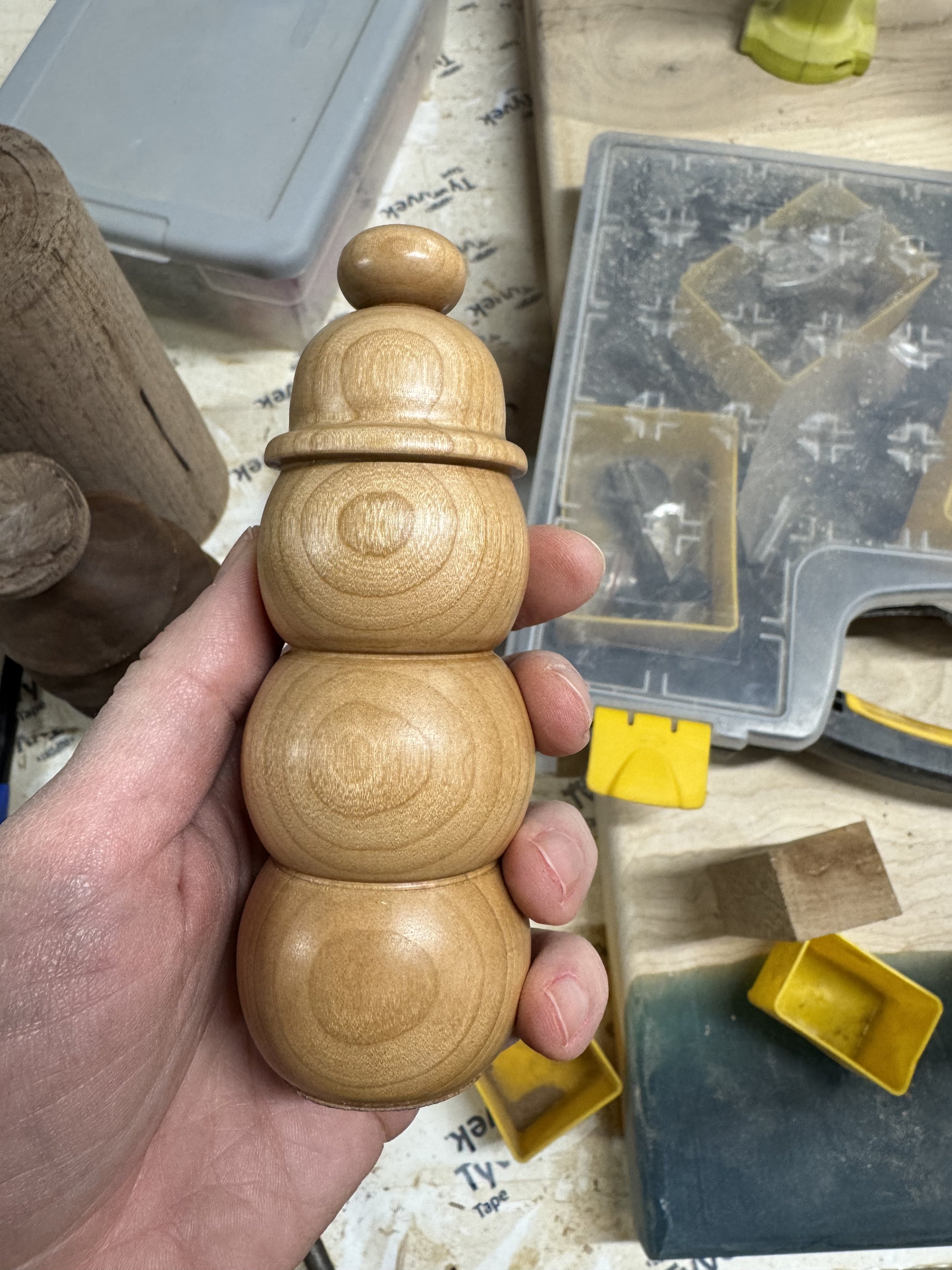 Hand holding a wooden snowman-shaped craft piece in a woodworking workshop, with plastic storage containers and woodworking tools in the background.