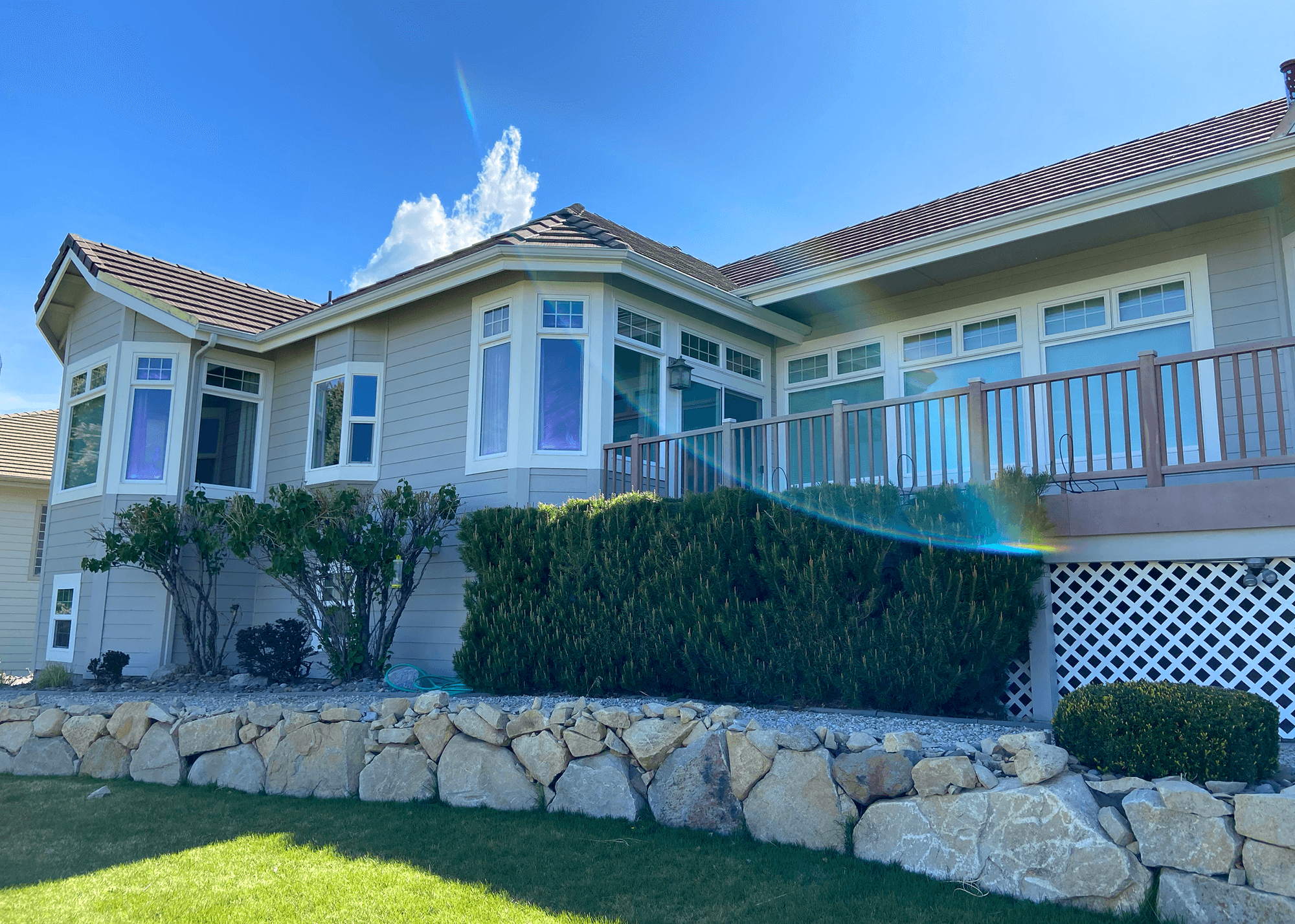 Back of a suburban house with multiple windows, a balcony, and a stone retaining wall in a sunny day.