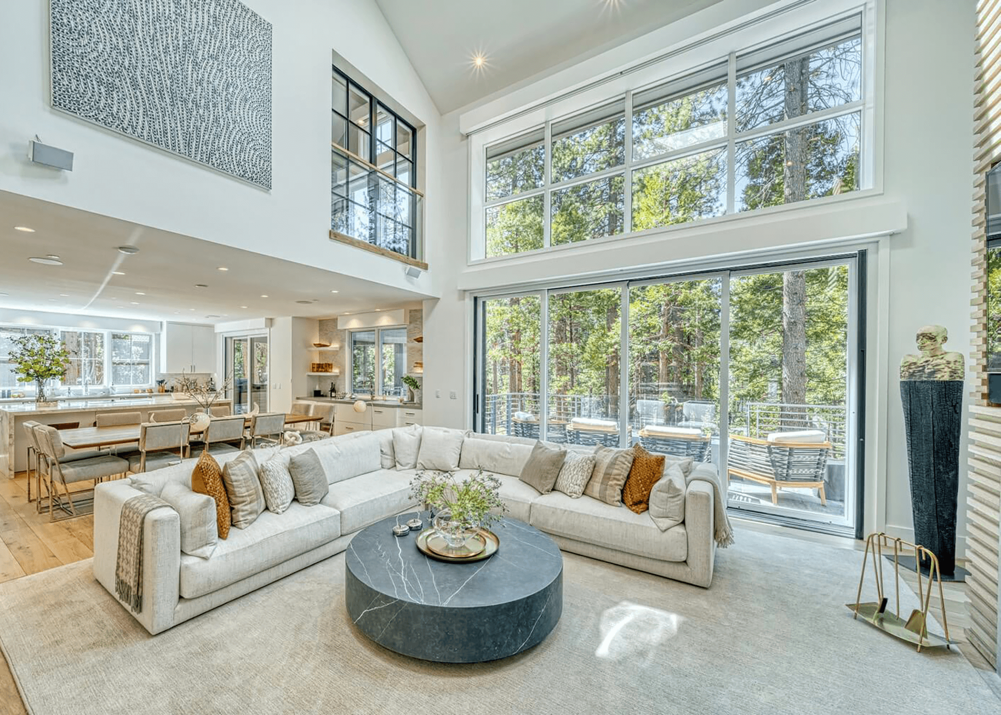 Open living room with large windows overlooking woods, white sectional sofa with beige and brown pillows, round black marble coffee table, dining area with chairs, contemporary decor, and wood flooring.