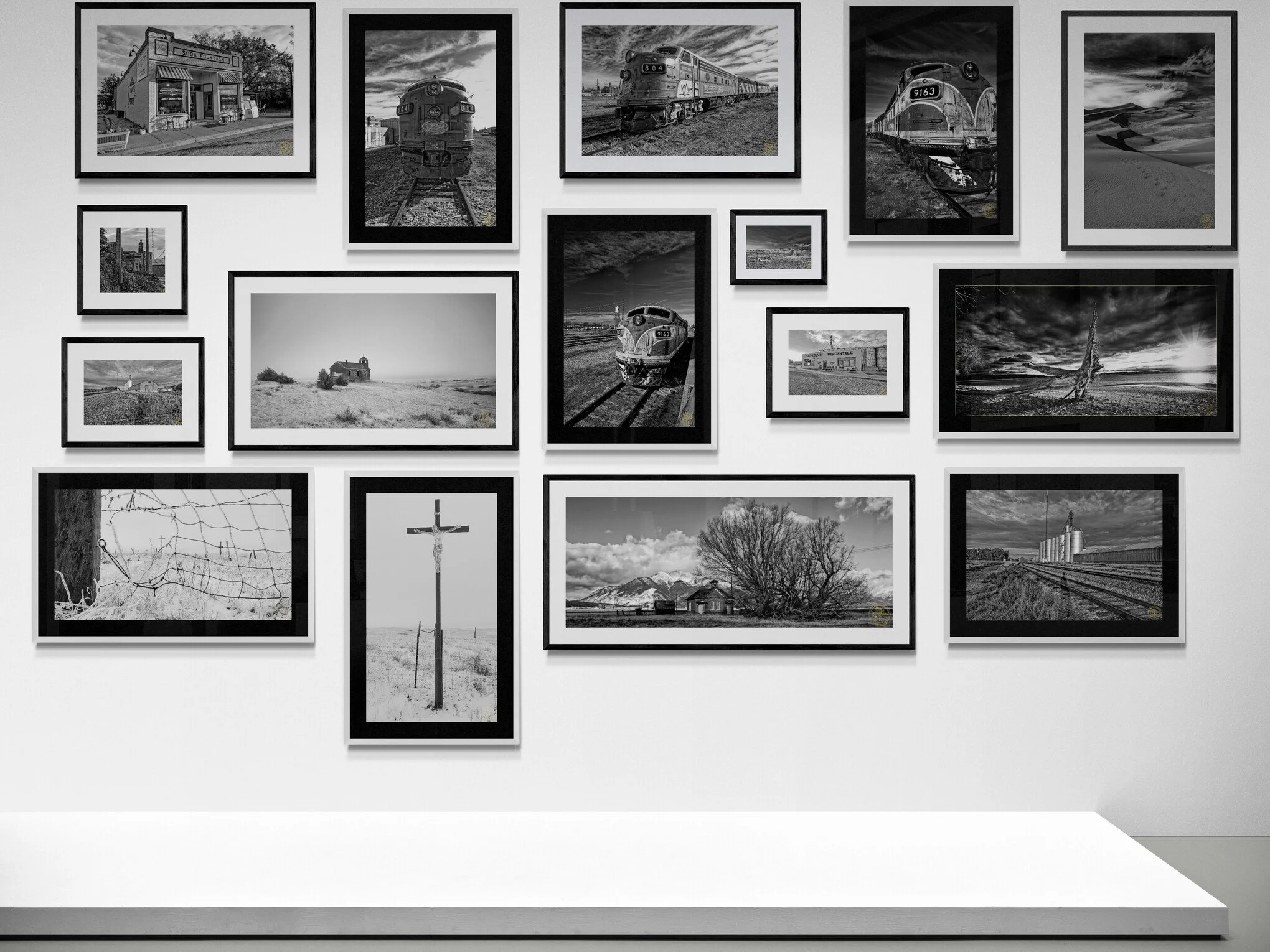 A collection of black and white photographs displayed on a gallery wall, featuring trains, landscapes, buildings, and natural scenes.