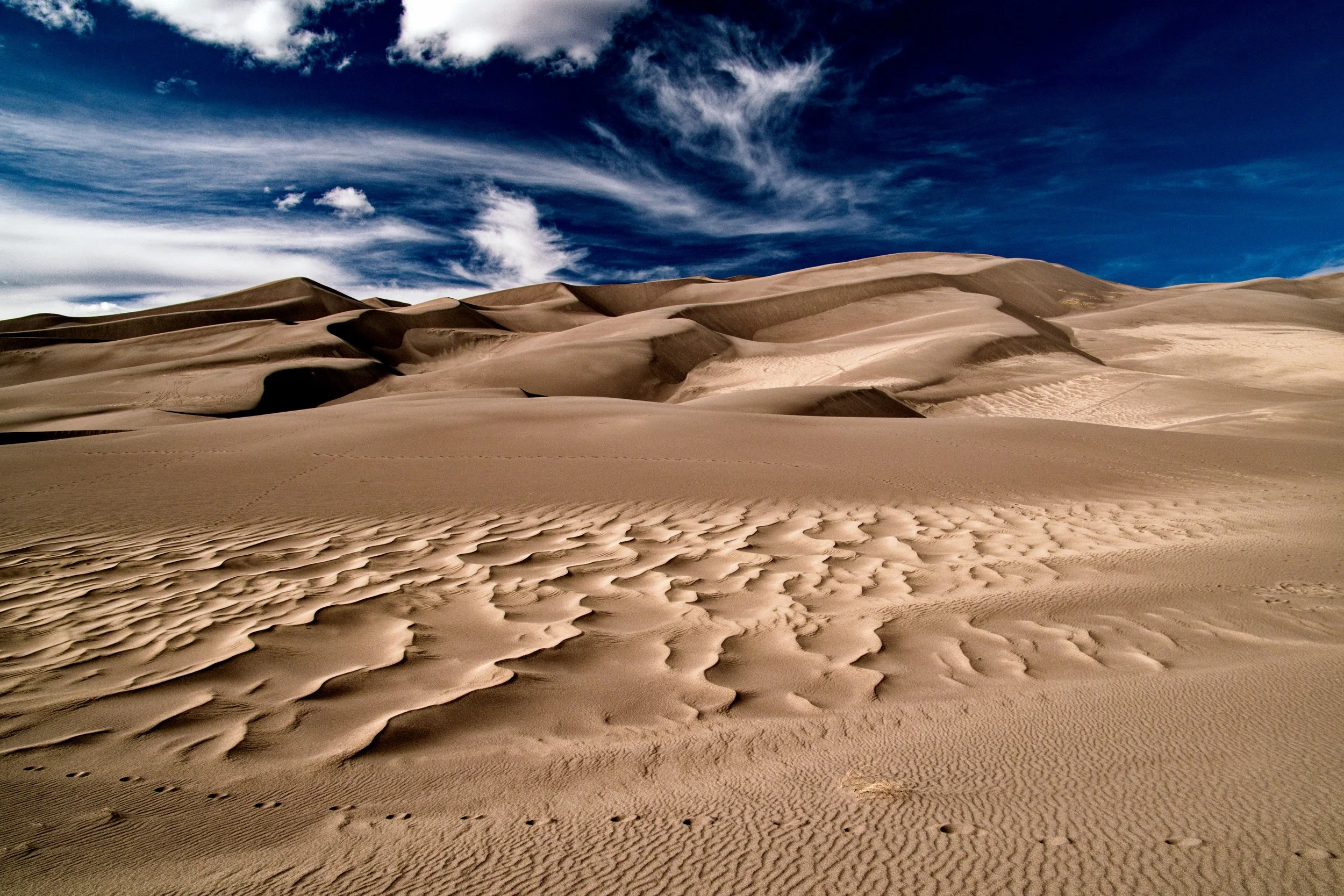 Wind-sculpted sand patterns leading toward towering dunes beneath dramatic clouds at Great Sand Dunes National Park in Colorado.