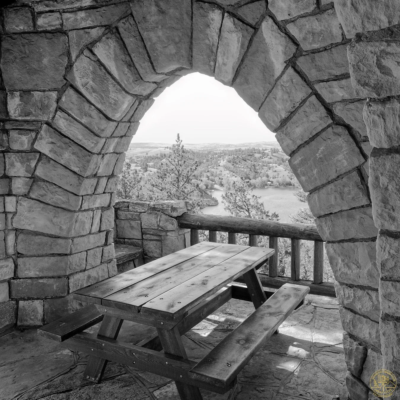 Black and white photo of a stone lookout tower with a wooden picnic table and railing, overlooking a landscape of trees, a river, and hills in the distance.