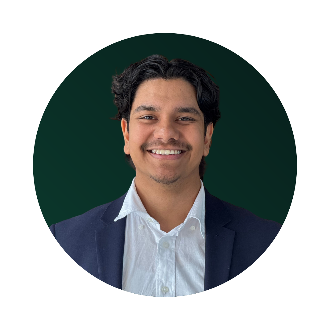 Headshot of a man with dark hair smiling, wearing a white shirt and navy blazer against a dark green background.