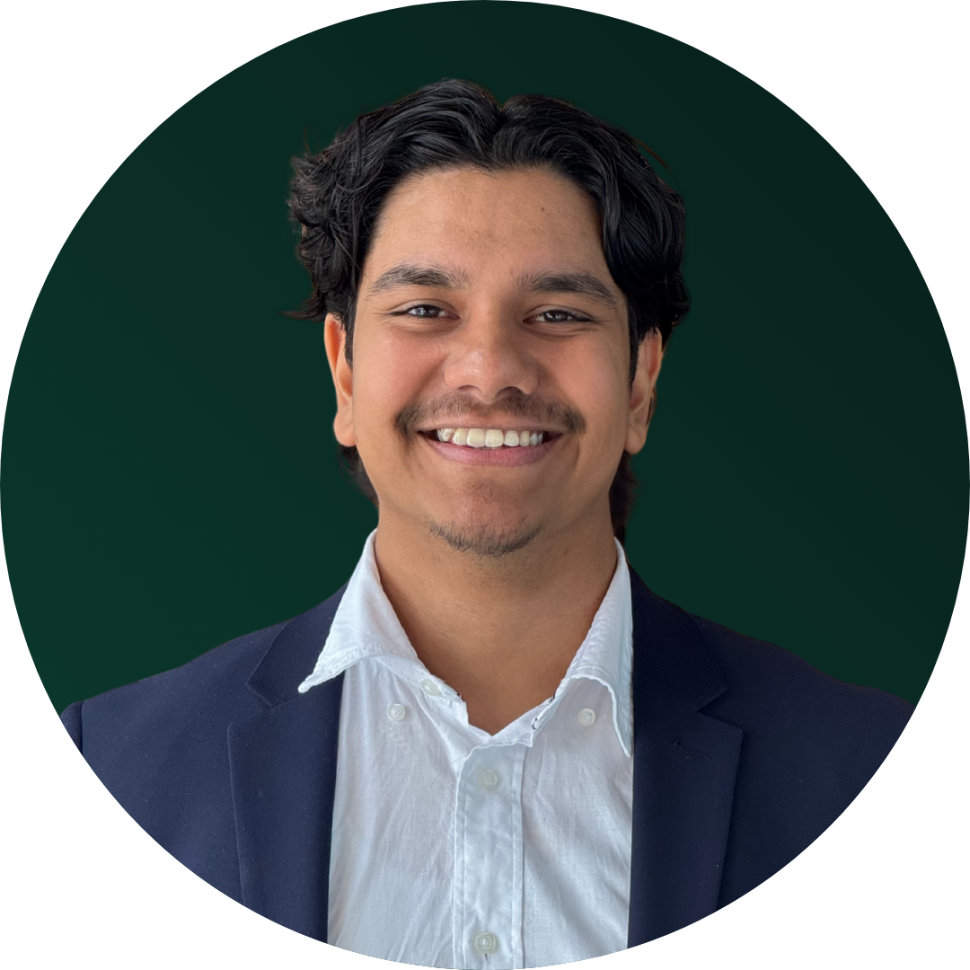 Headshot of a man with dark hair smiling, wearing a white shirt and navy blazer against a dark green background.