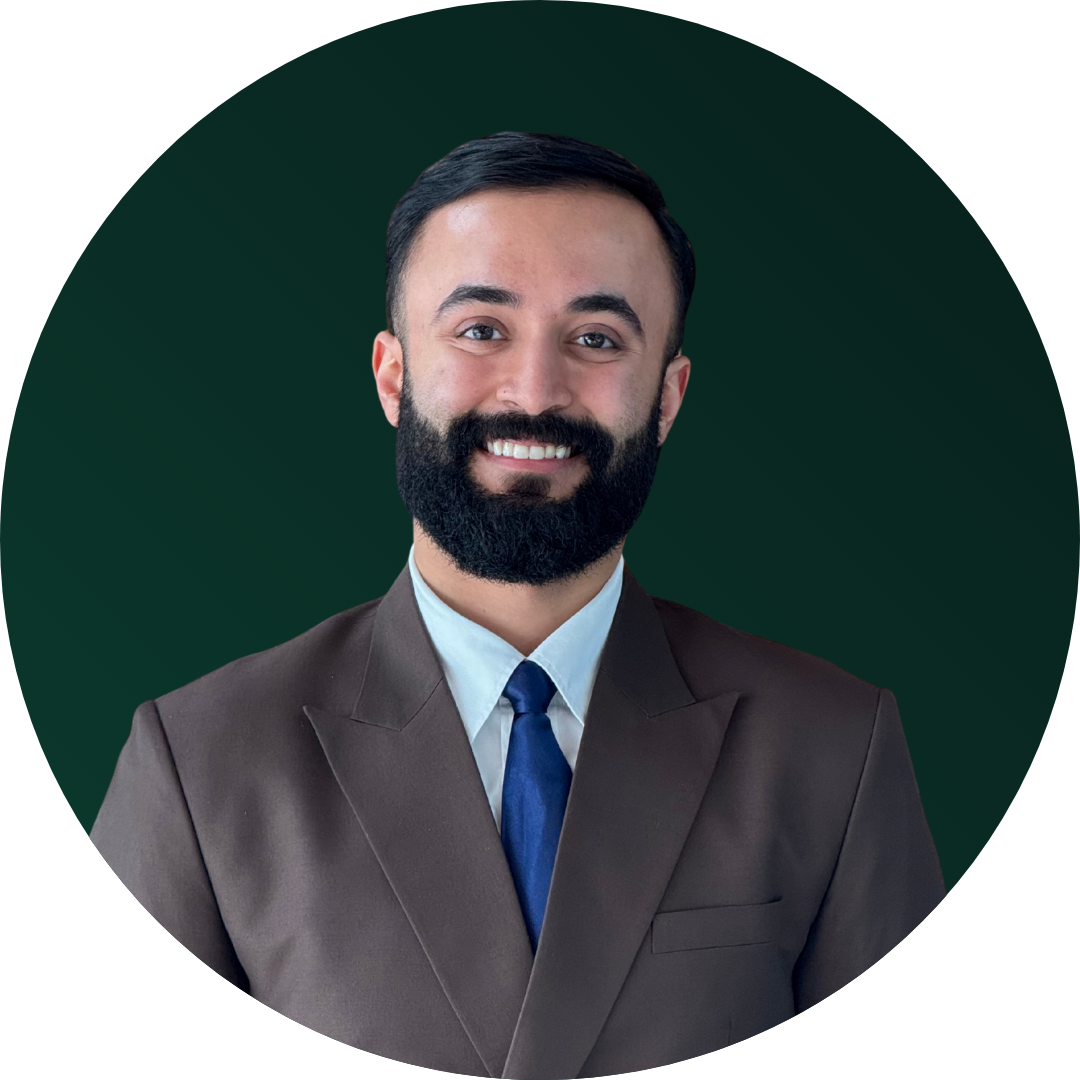 Headshot of a man with dark hair, a full beard, wearing a brown suit, white shirt, and blue tie, smiling against a dark green background.