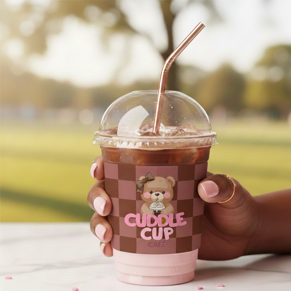 Hand holding a cup of iced chocolate with a dome lid and a pink straw, featuring a bear logo and the words "Cuddle Cup Café" on the cup, outdoors with a blurred park background.