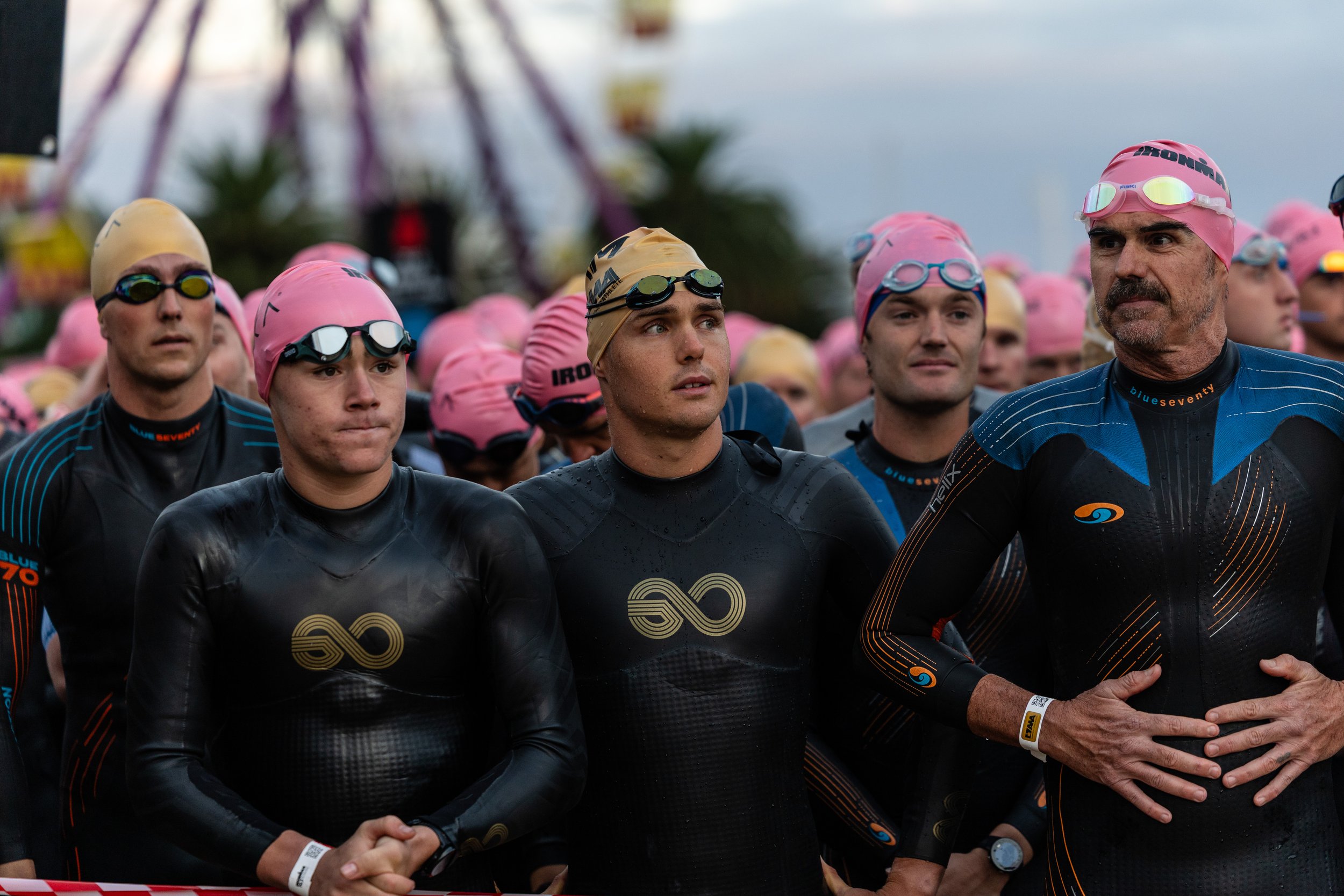Group of male triathletes in wetsuits and pink swimming caps at the starting line of a race, outdoors.