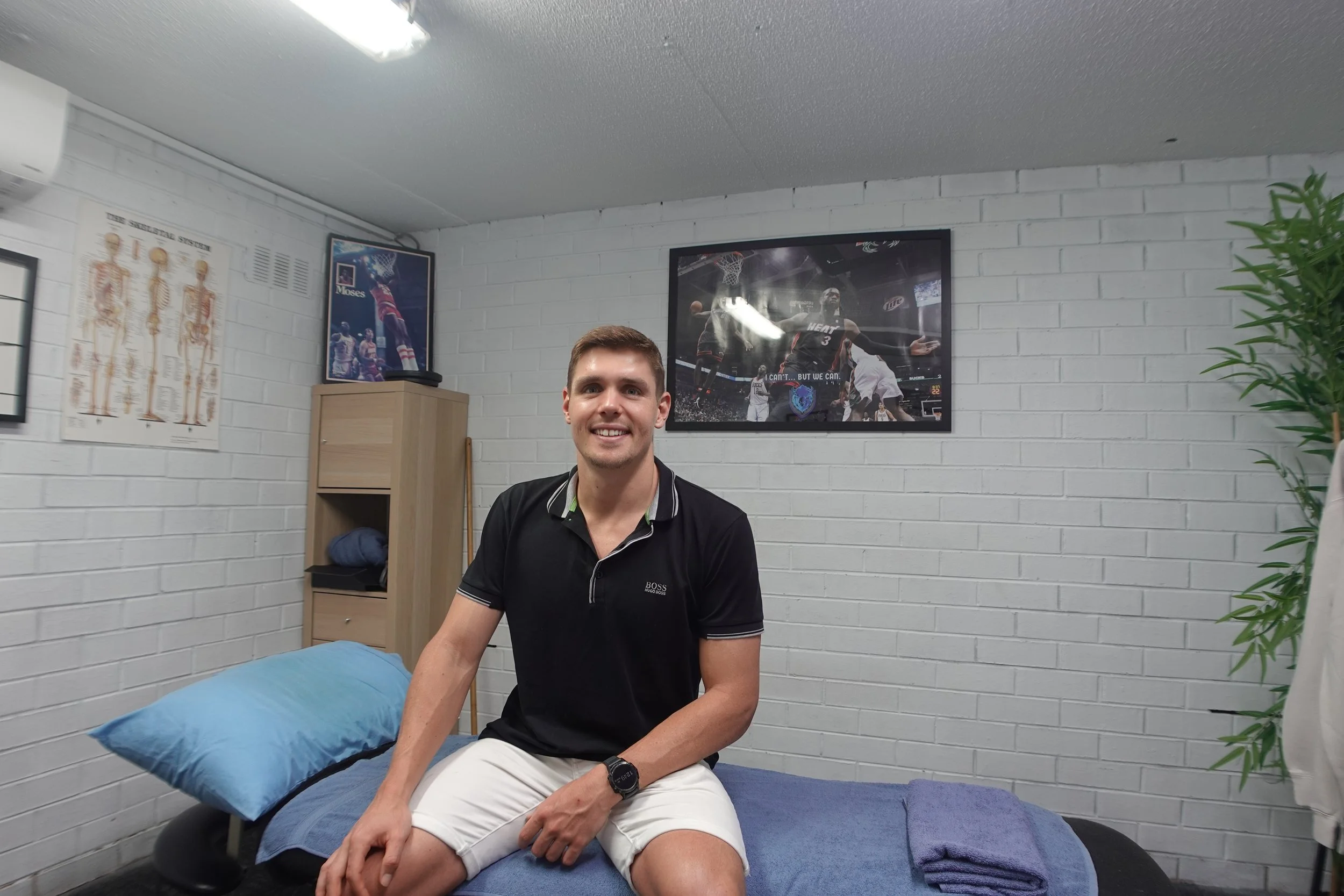 A smiling young man sitting on an examination table in a chiropractor's office, with framed sports posters and a potted plant on white brick walls.