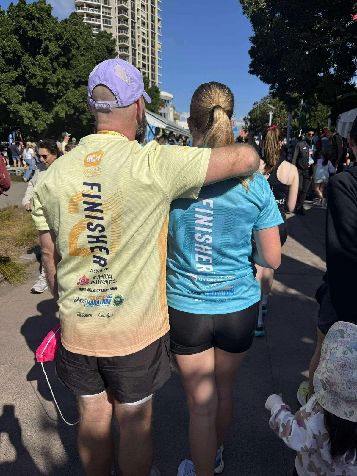 People at a marathon finish line, two individuals in colorful 'FINISHER' shirts walking together with their arms around each other's shoulders, surrounded by other runners and spectators.