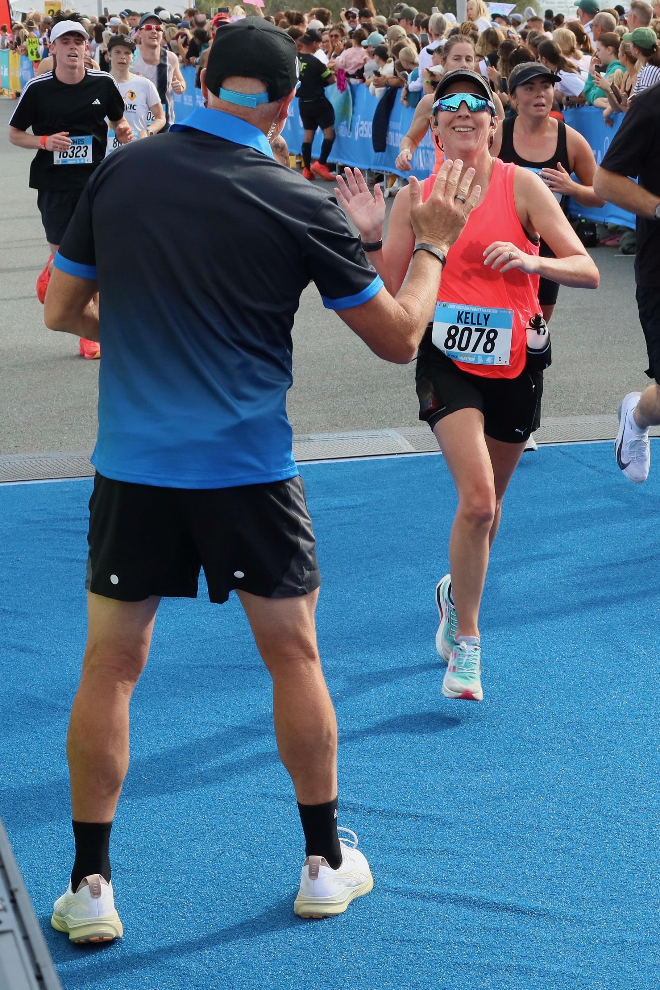 A woman runner high-fives a man at a marathon finish line, with many spectators and other runners in the background.