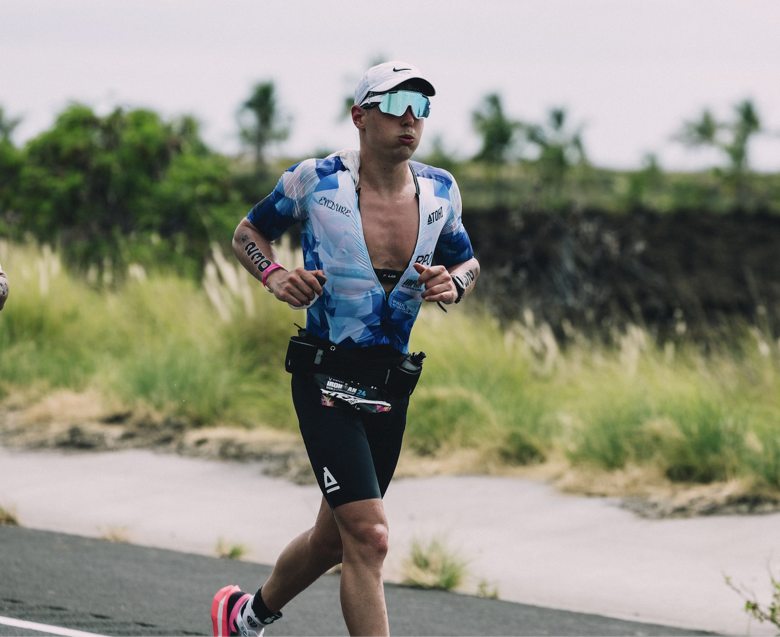 Male triathlete running outdoors in a blue and white sports outfit with sunglasses and a white cap.