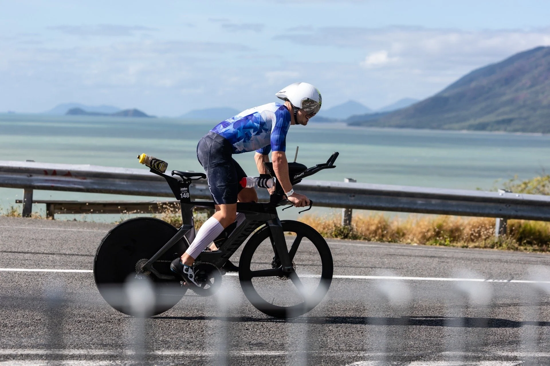 A person wearing a blue cycling jersey, black shorts, and a white helmet riding a black time trial bicycle along a scenic highway with mountains and water in the background.